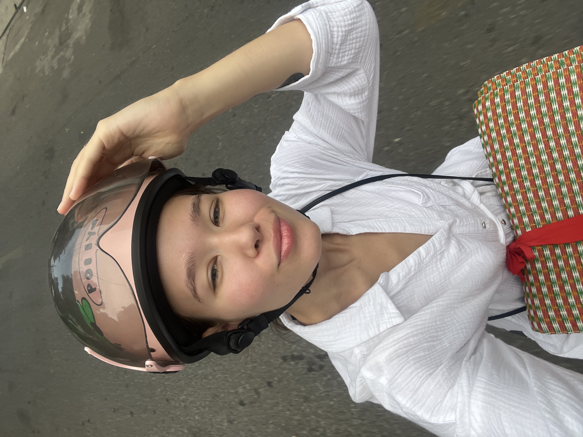 A young woman is depicted from an overhead perspective, likely on a scooter or motorcycle, in what appears to be a street setting in Nha Trang Ward, Vietnam. She is wearing a textured white long-sleeved shirt and a pink and black helmet with a clear visor. Her left hand is raised to her temple, lightly touching the helmet's edge.
The woman is looking directly at the camera with a subtle, content smile. Her cheeks show a natural flush. On the side of her pink helmet, partial text that reads "POLI" or "PUI" is visible, along with a green graphic, possibly a leaf or abstract design.
To her right, a woven bag or basket is seen, featuring a vibrant geometric pattern of red, green, and white stripes, secured with a red fabric tie. The background consists of a dark grey asphalt surface, suggesting a road or paved area. The lighting indicates it is daytime. The scene conveys a casual, everyday activity in an urban or semi-urban environment.