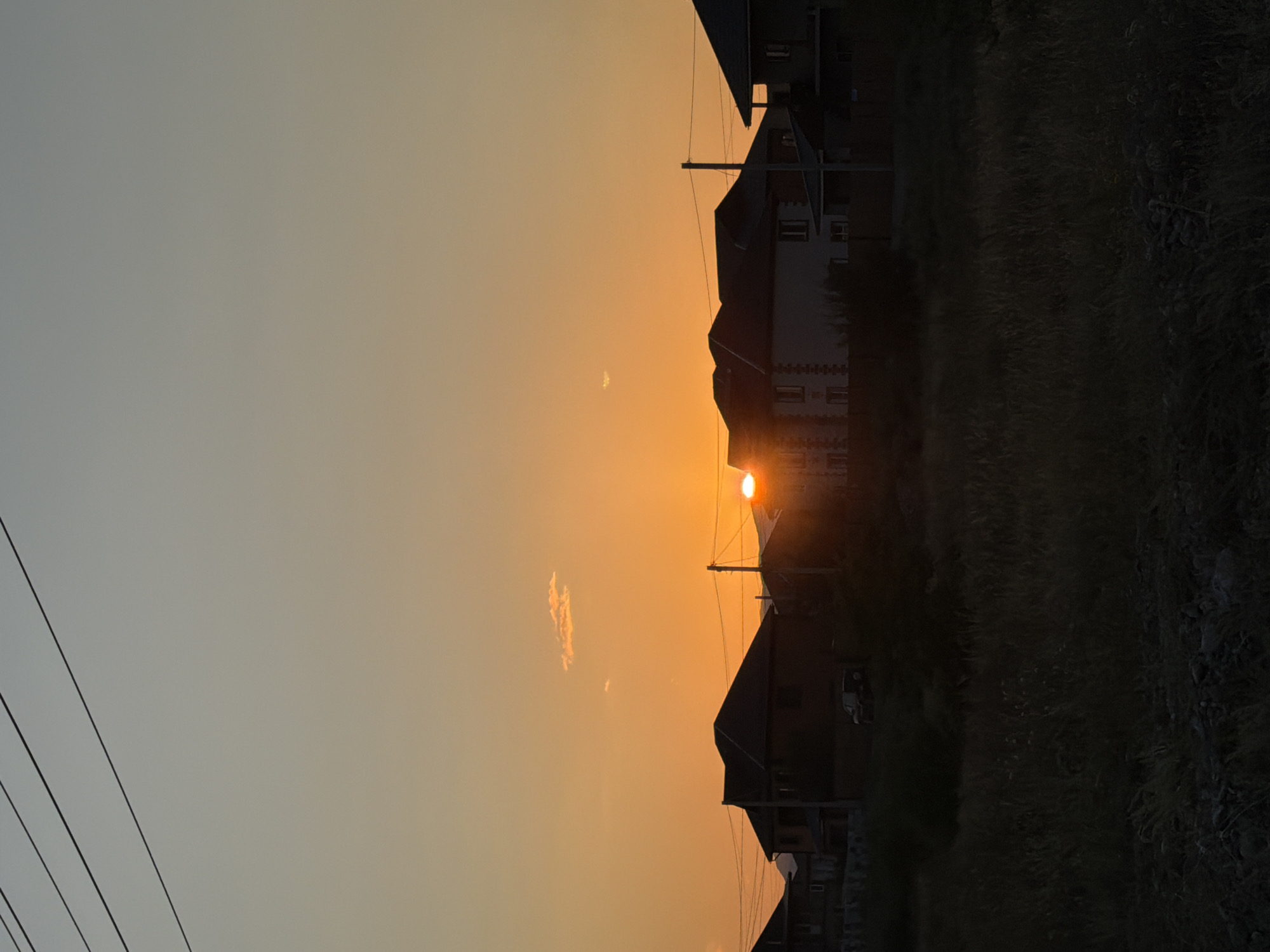 The image shows a residential area at sunset, likely in Kyzyl, Russia, due to the distinctive architecture and landscape. Silhouetted against a vibrant orange sky are several two-story houses with dark-tiled roofs. A few power lines crisscross the upper portion of the frame, and a single small cloud is visible. The foreground is dominated by dark, dry grass and some rocks, suggesting a rural or semi-rural setting on the outskirts of the city. The sun is partially visible on the right side of the image, casting a warm glow and creating silhouettes of the buildings. A dark-colored SUV is parked in front of one of the houses. The overall mood is peaceful and serene, typical of a sunset in a developing area.