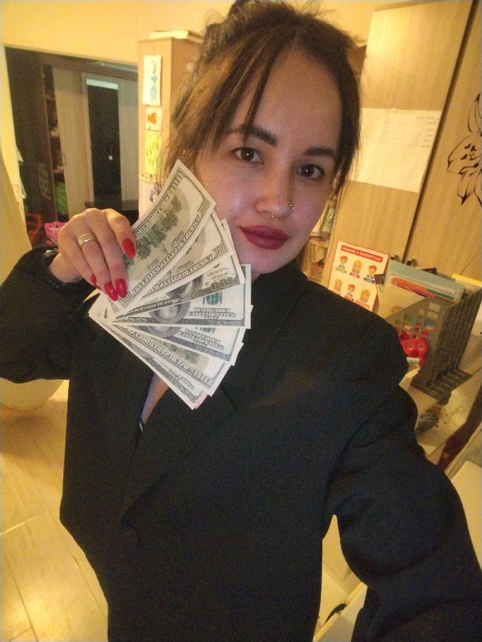 A woman in a black blazer holds a fan of US $100 bills. She has brown hair, red lipstick, a nose ring, and a ring on her left ring finger. The background shows a room interior with a door, a cabinet with drawings and papers attached, and miscellaneous items on a table. The lighting is a bit dim, with warm tones. The photograph has a selfie perspective, and the woman appears to be the main subject.