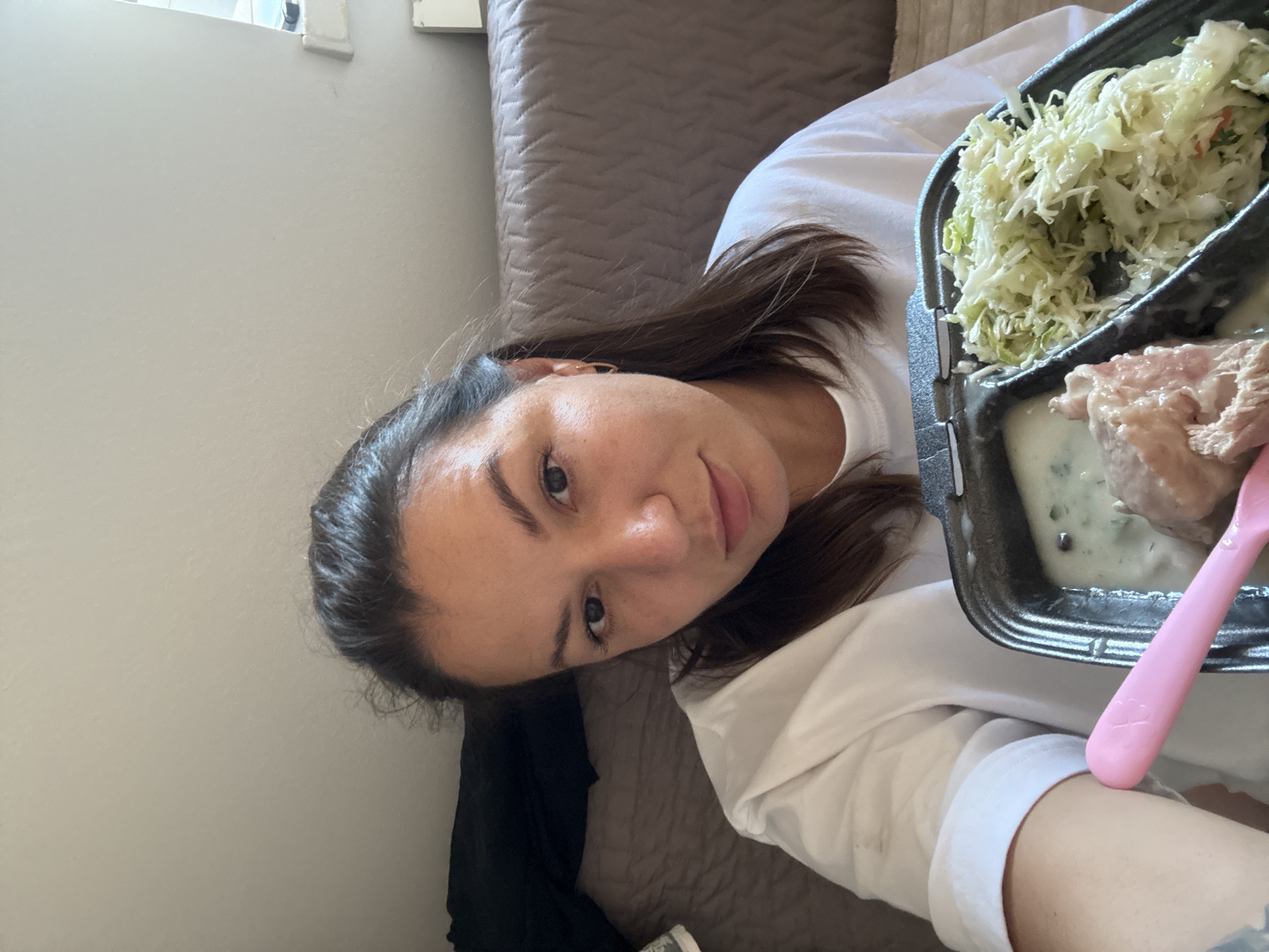 A young adult female, with brown hair pulled back and dark eyes, is holding a black plastic take-out container with two compartments, as she looks at the camera. The container holds a portion of what appears to be shredded coleslaw in one compartment and shredded meat with a creamy sauce in the other. A pink plastic utensil rests across the front of the container. The woman is wearing a white t-shirt. The background consists of a textured brown couch and a beige wall. The scene appears to be indoors. There is no indication of the city of Września, Poland.