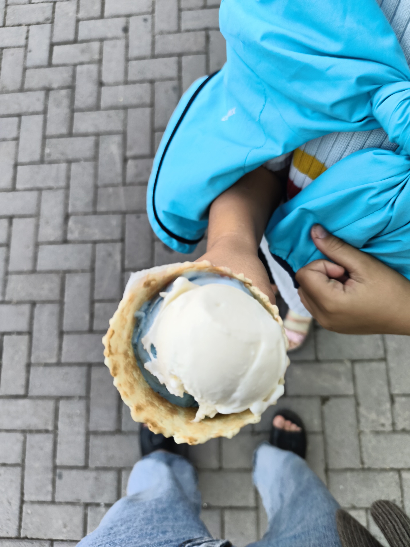 An overhead shot shows a person in light blue jeans and black sandals holding a waffle cone with two scoops of ice cream. The ice cream appears to be vanilla and blue raspberry. Another person, wearing a bright blue jacket over a striped shirt, is partially visible in the frame. They are standing on a brick-paved walkway, likely in an outdoor urban setting. The image lacks specific indicators of the city of Gorno-Altaysk, Russia.