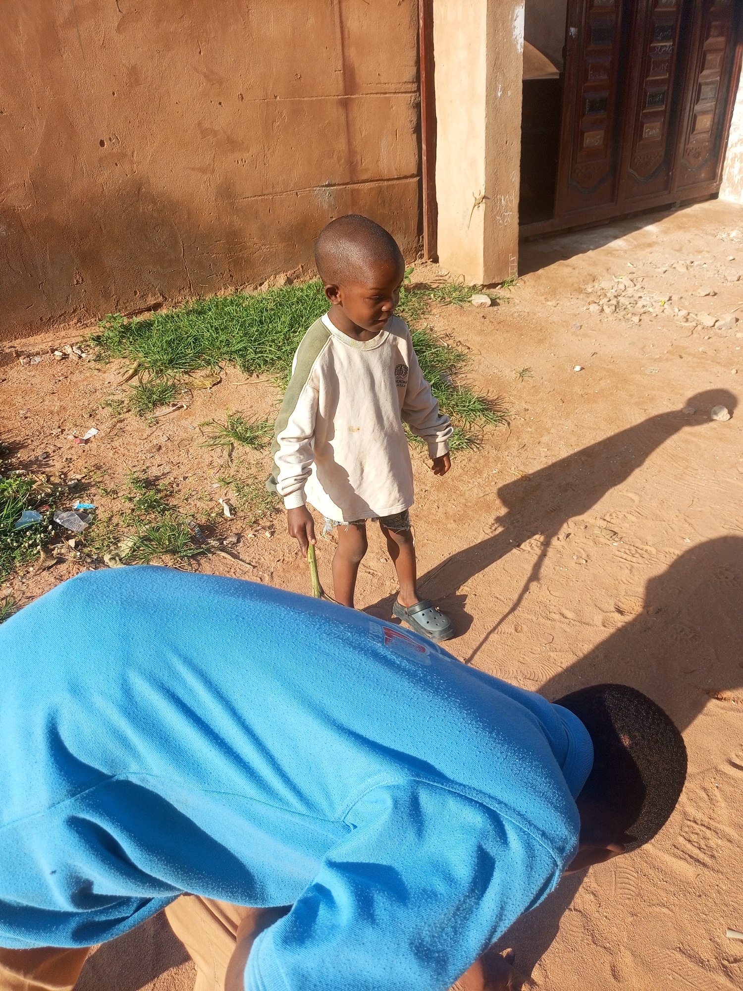 An exterior daytime shot shows a young boy standing in a sandy area with sparse green grass. The boy, facing away from the camera, wears a light-colored long-sleeved shirt and shorts with dark clogs. He holds a thin green stalk in his right hand. In the foreground, the back of another person wearing a bright blue long-sleeved shirt is visible, facing away and bending forward. The background features a textured, reddish-brown wall with a vertical pillar and a dark wooden door to the right, suggesting a residential or public building in Tempure, Nigeria. The ground is sandy with small debris, and shadows indicate a bright, sunny day.