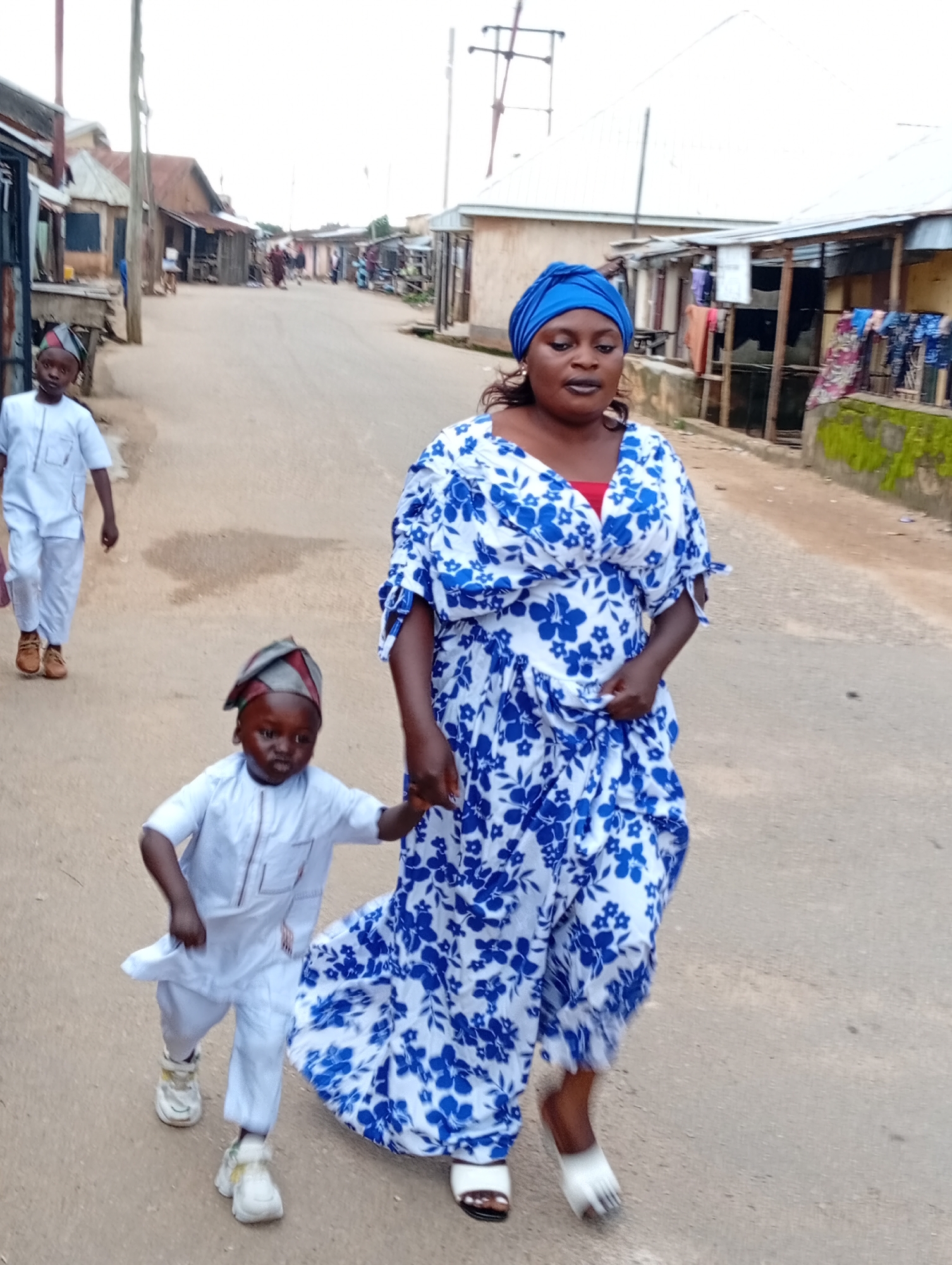 A medium shot photograph captures a street scene in Jos, Nigeria, where a woman in a blue and white floral dress and a blue headscarf walks hand-in-hand with a young child wearing a white outfit and a patterned hat. Another child, also in a white outfit, walks behind them to the left of the frame. The background shows a street lined with buildings that appear to be shops or homes, with some laundry hanging outside. The ground is a dusty, paved surface. The lighting suggests it is daytime, possibly overcast.