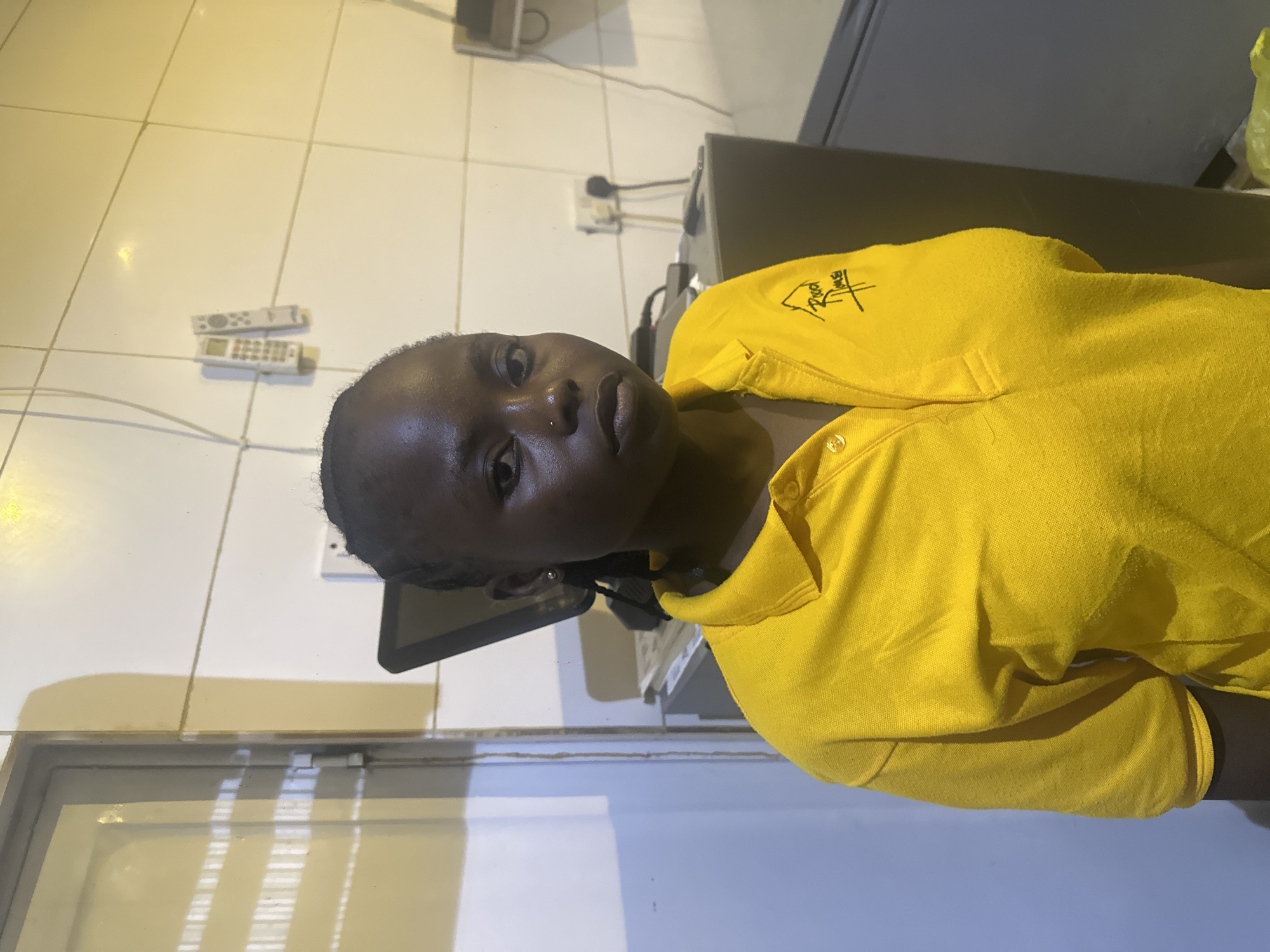 A young Black woman with dark braided hair and a nose ring is pictured from the chest up, facing the camera. She wears a bright yellow polo shirt with a logo on the left chest. The background suggests an indoor setting with a white tiled wall, a closed metal door on the left, and various electrical outlets and devices on the wall behind her. The lighting is warm and casts shadows. The scene appears to be within a commercial or administrative space.
