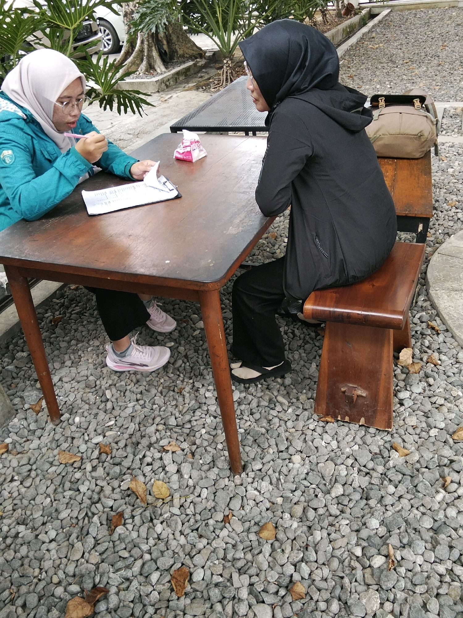Two individuals are seated at a rustic wooden table outdoors, which is situated on a gravel surface. The person on the left, wearing a teal jacket and a white hijab, is looking down at papers on a clipboard and appears to be writing or marking them with a pink pen. This individual is wearing glasses and pink sneakers. The person on the right is wearing a black hijab and a black jacket, and is seated on a wooden bench. Their body is angled away from the camera. A beige backpack rests on the bench beside them. The surrounding environment includes foliage and trees, and a parked car is partially visible in the background. The setting suggests an outdoor cafe or seating area in the city of Binorong, Indonesia.