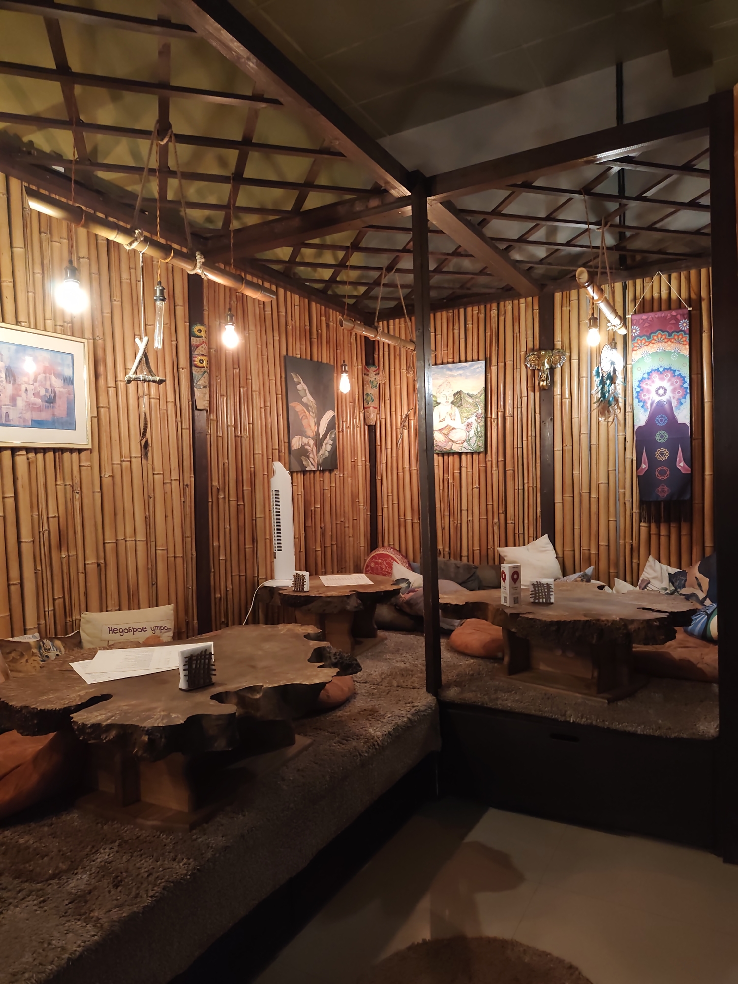 The image depicts the interior of a dimly lit, bohemian-style cafe or lounge area, likely in Sochi, Russia, with walls and ceiling constructed from bamboo. The seating consists of low, rustic tables made from natural wood slabs, set on thick, textured mats, with numerous scattered cushions. Hanging pendant lights illuminate the space, casting a warm glow. Several pieces of artwork adorn the bamboo walls, including a framed painting on the left, a depiction of a figure meditating in a landscape, and a vibrant tapestry featuring a chakra diagram and a silhouette of a person. Decorative items like dreamcatchers and a golden elephant figurine are also visible. A white standing fan is positioned near one of the tables. The overall atmosphere suggests a relaxed and possibly spiritual or artistic establishment.