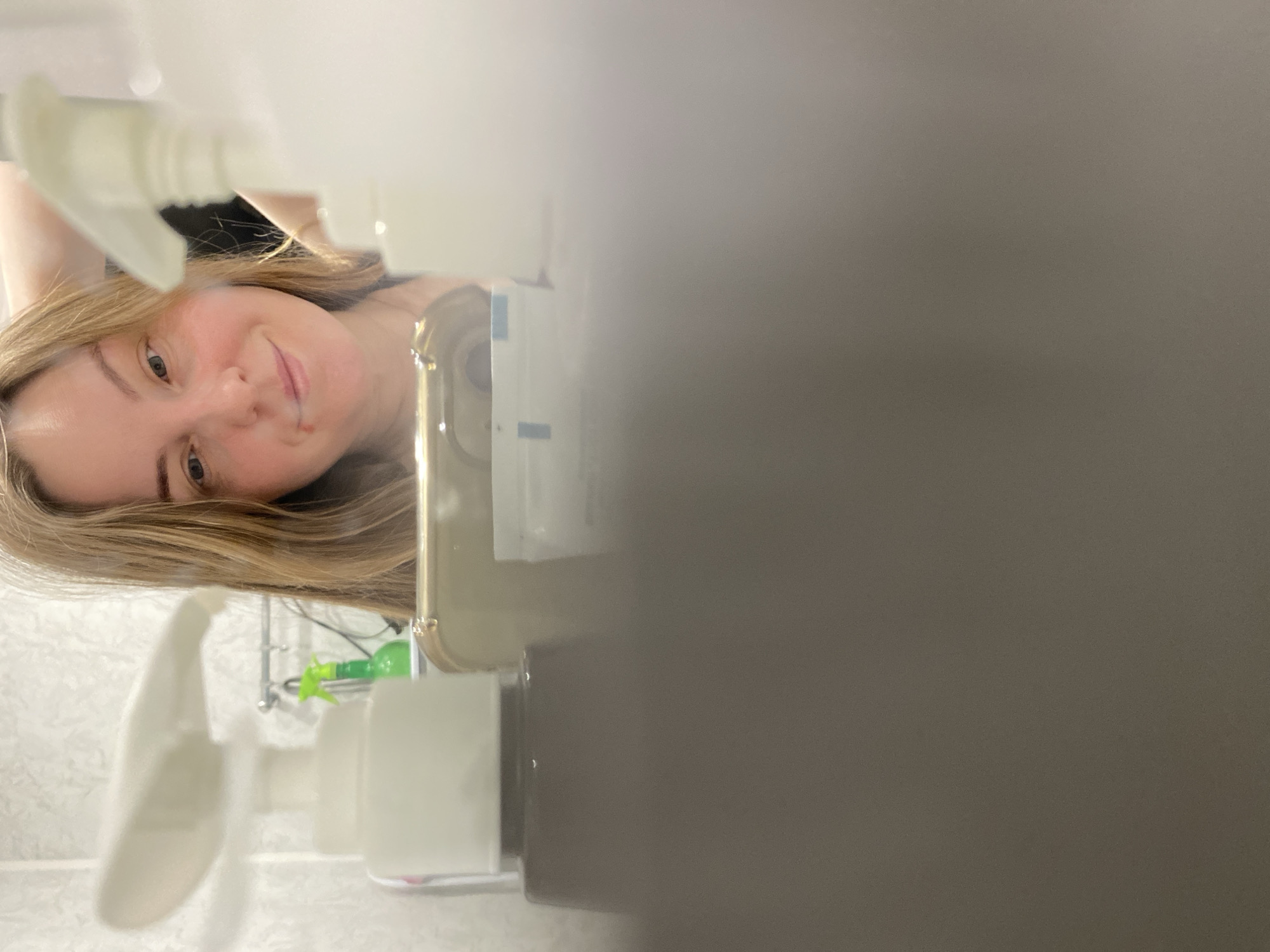 A woman with fair skin and blonde hair is visible in a partial selfie taken in a bathroom setting. Her face is positioned in the upper left quadrant. The lower left quadrant shows bathroom fixtures including a white tiled wall, a metal shower rack, and various white plastic bottles with dispensing pumps. A green spray bottle is also visible. The right half of the image is dominated by a blurry, dark grey or brown surface, obscuring details. The photo is likely taken with a phone camera, given the presence of the phone edge visible along the right side of her face. The scene appears to be inside a domestic bathroom setting, possibly in an apartment or house. There is no verifiable connection to the city of Noviy, Russia, evident from the provided image.