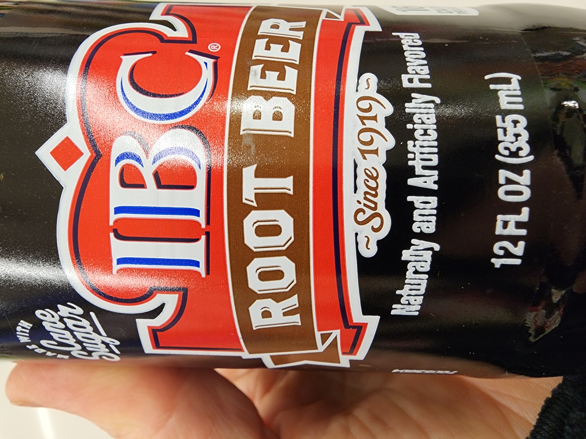 The image is a close-up, eye-level, outdoor shot of a person holding an IBC Root Beer bottle. The bottle is black with a red and white label. The prominent text on the label includes "IBC," "ROOT BEER," "Since 1919," "Naturally and Artificially Flavored," and "12 FL OZ (355 mL)." The person's hand, which is holding the bottle, is visible on the left side of the frame. The background is blurred and appears to be an indoor setting. The image was taken in Camp Hill, United States of America.