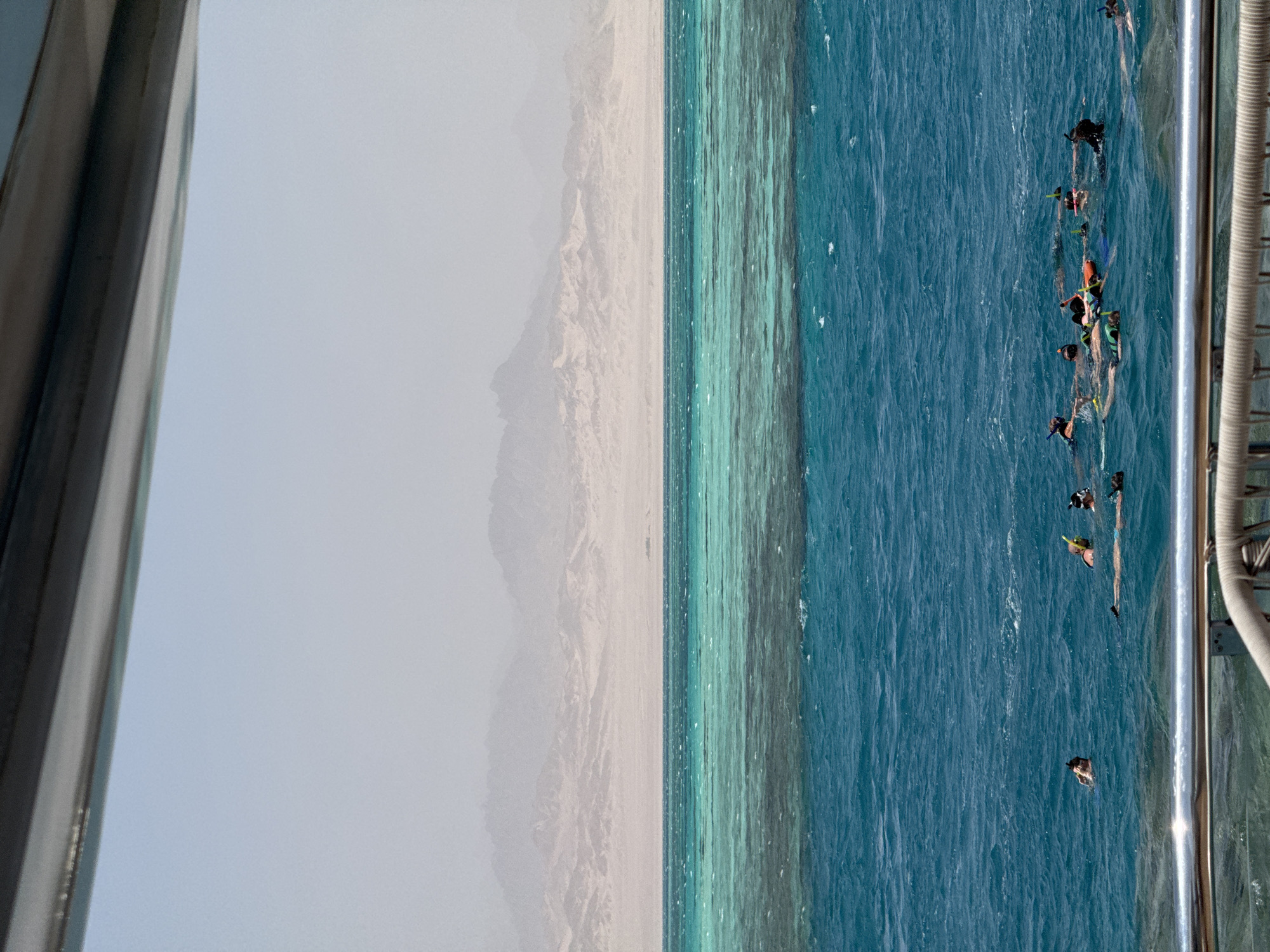 The image shows a group of snorkelers in the turquoise waters of the Red Sea, with a barren, mountainous landscape in the background. The snorkelers, wearing masks and snorkels, are scattered across the water, some swimming and others appearing to be observing something below the surface. The water exhibits a distinct gradient of color, from a lighter, almost translucent turquoise near the shore to a deeper, richer blue further out. The distant mountains are shrouded in a hazy atmosphere, suggesting a warm, arid climate. The foreground features the edge of a boat or pier with a metal railing, indicating that the snorkelers may have arrived by boat. The scene captures a typical tourist activity in a coastal region of Egypt, likely a popular snorkeling spot.