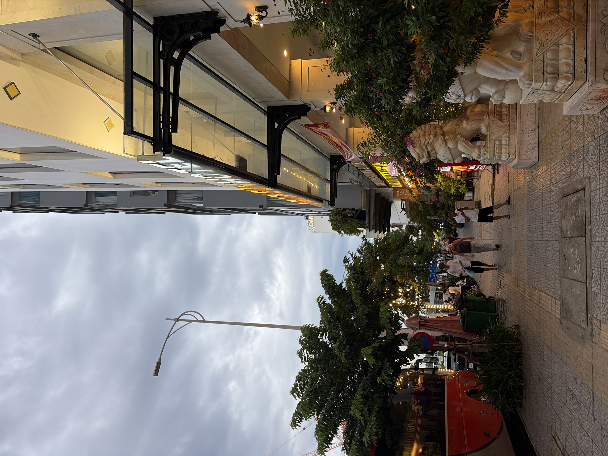 The image shows a sidewalk in front of a modern building in Đà Nẵng, Vietnam. A red tour bus is parked on the left side of the street. Several people are walking on the sidewalk, some are talking to each other, while others are walking alone. There are trees and streetlights along the sidewalk, and decorative lion statues are placed in front of the building. The sky is cloudy, indicating that it might be evening or a cloudy day. The building appears to be a hotel or a commercial establishment with a glass awning over the entrance. The overall scene depicts a typical urban environment in Vietnam.