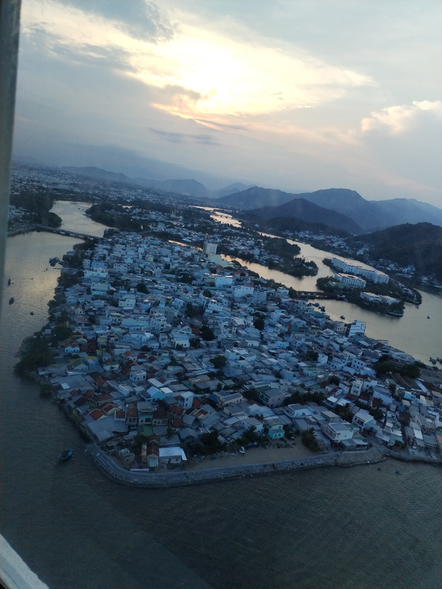 The image presents an aerial view of Bac Nha Trang Ward, Vietnam, taken during the daytime with a cloudy sky. The sun is partially obscured. A river winds through the landscape, surrounding an island densely packed with buildings. Mountains are visible in the background. The river has several small islands or sandbars. The image was taken from an elevated perspective, possibly from an aircraft or tall structure.