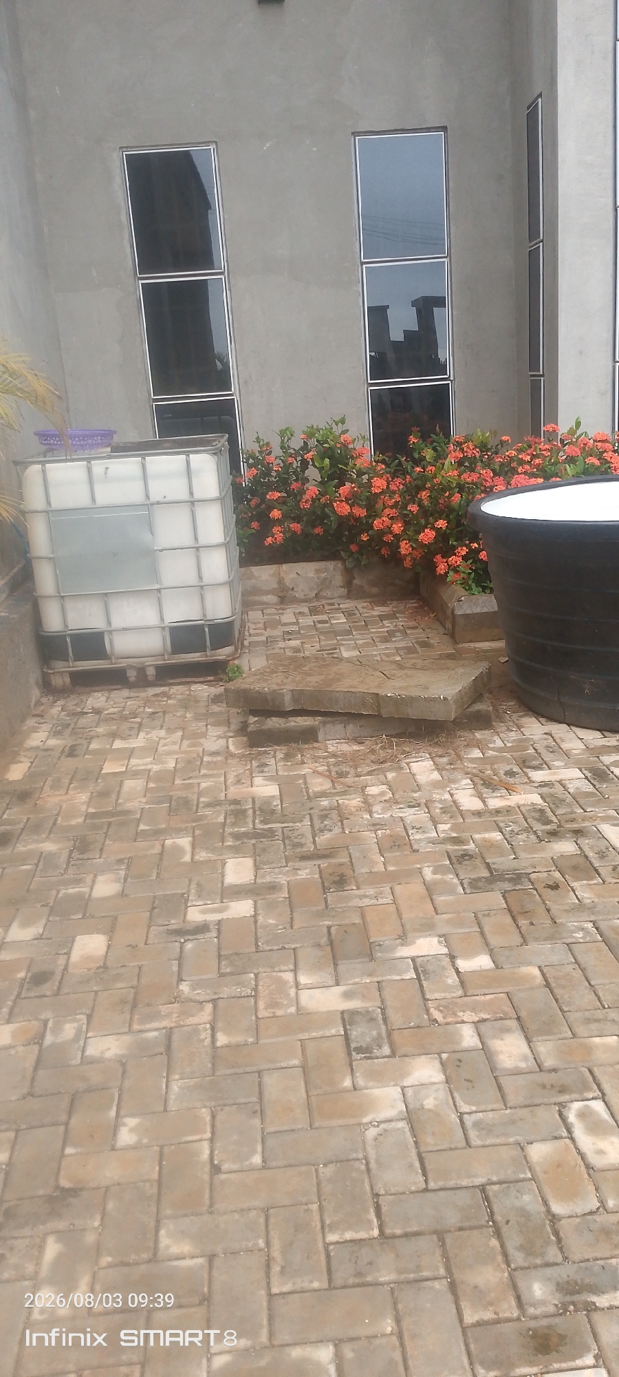 The image shows a paved courtyard area with a building in the background. The building has a gray, textured wall with several tall, narrow windows. In front of the building, there is a large white cube-shaped tank with a metal cage around it, possibly an IBC tote. To the right of the tank, there is a bed of flowering plants with small orange flowers. A large black cylindrical tank is visible on the right side of the image. The courtyard pavement is made of interlocking brick pavers laid in a herringbone pattern. There are some flat stone slabs on the ground in front of the flower bed. The overall scene appears to be an outdoor storage or utility area, possibly associated with a residential or commercial building in Kakau, Nigeria. The bottom left corner of the image shows a timestamp "2026/08/03 09:39" and "Infinix SMART8".