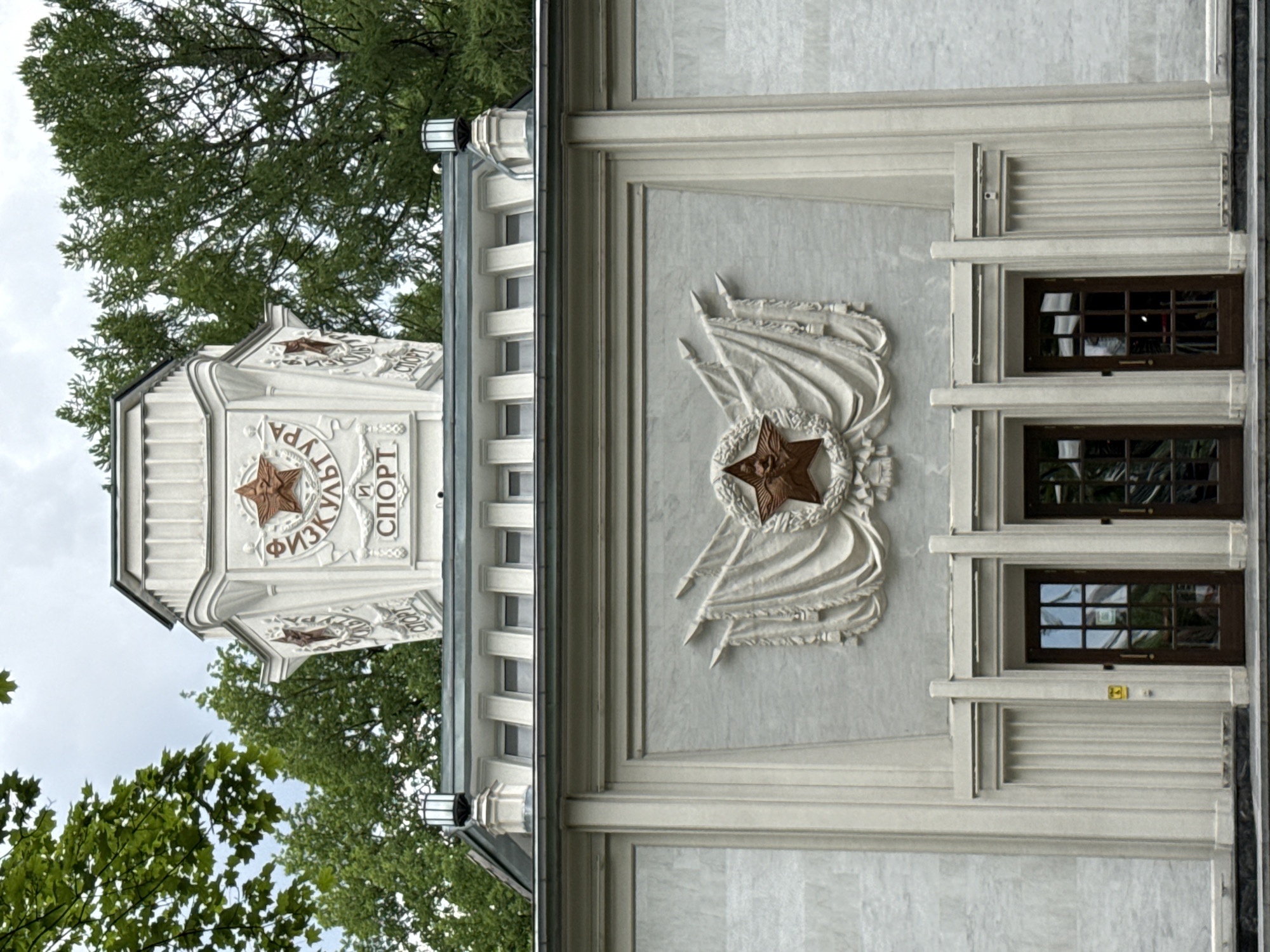 The image shows the facade of a building with Soviet-era architectural elements. The building is likely located in Moscow, Russia, possibly within the VDNKh (Exhibition of Achievements of National Economy) park, given the style of architecture and the prominent Soviet symbols.
The central feature of the facade is a large bas-relief depicting a red star emblem surrounded by laurel leaves and draped fabric, symbolizing Soviet achievement and victory. Above this, on a tiered structure, are inscriptions in Russian. The upper part of the building features a cupola-like structure with more Soviet star emblems and inscriptions, reading "ФИЗКУЛЬТУРА" (Physical Culture) and "СПОРТ" (Sport).
The facade is made of light-colored stone or marble, with fluted columns and rectangular windows. There are three dark wooden doors with glass panes at the ground level. The building is flanked by trees and the sky is overcast. The overall impression is one of monumental Soviet architecture, emphasizing themes of physical culture and national achievement. There are no people visible in the image.