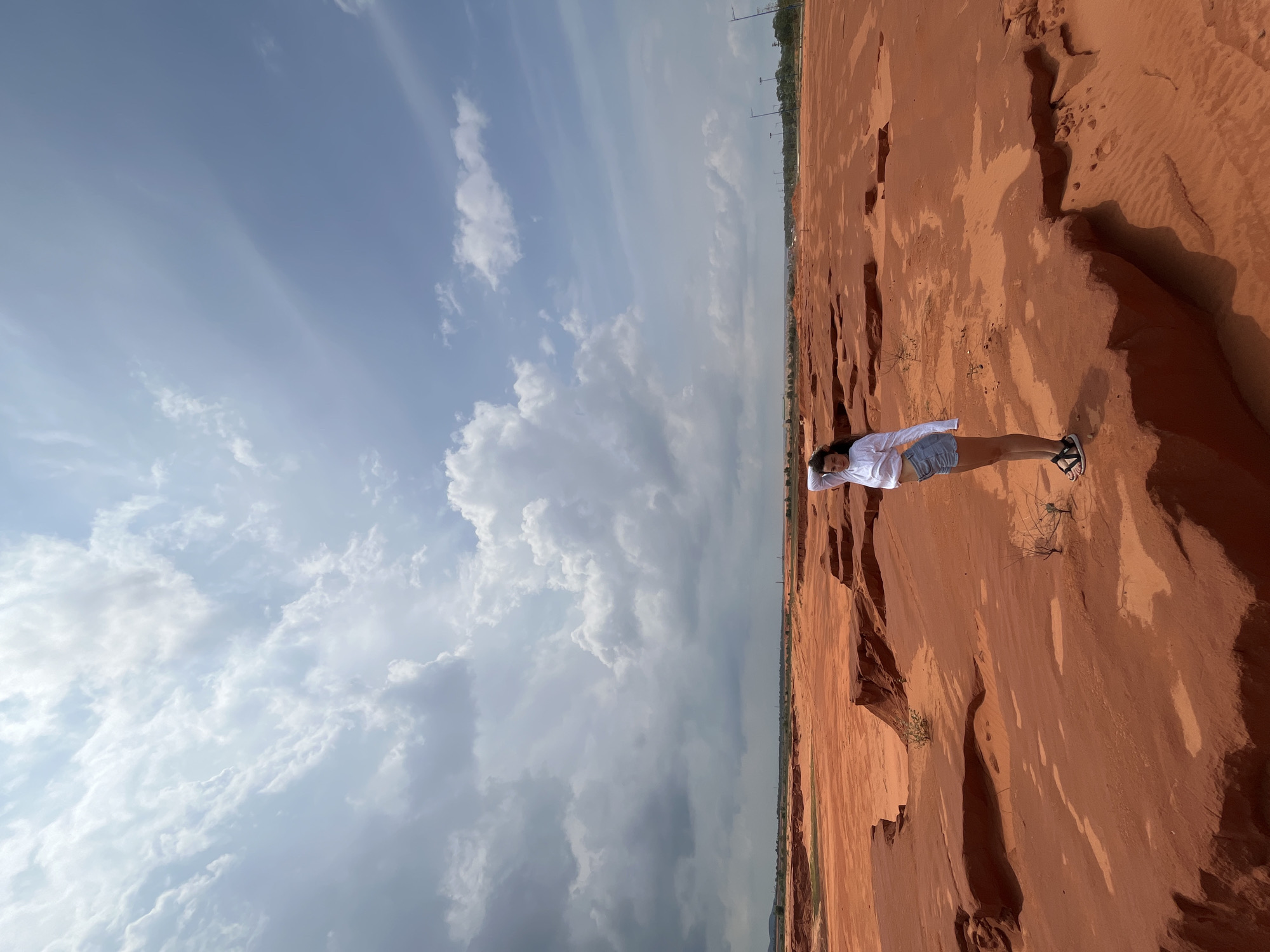 A young woman with dark curly hair stands on a ledge of red sand in Mui Ne, Vietnam. She is wearing a white long-sleeved shirt and light blue denim shorts, with her right hand raised to her head. The landscape is a vast expanse of red sand dunes with eroded formations and scattered dry vegetation. The sky above is a mix of blue and dramatic, dark clouds. In the distance, a line of trees and utility poles mark the edge of the desert.