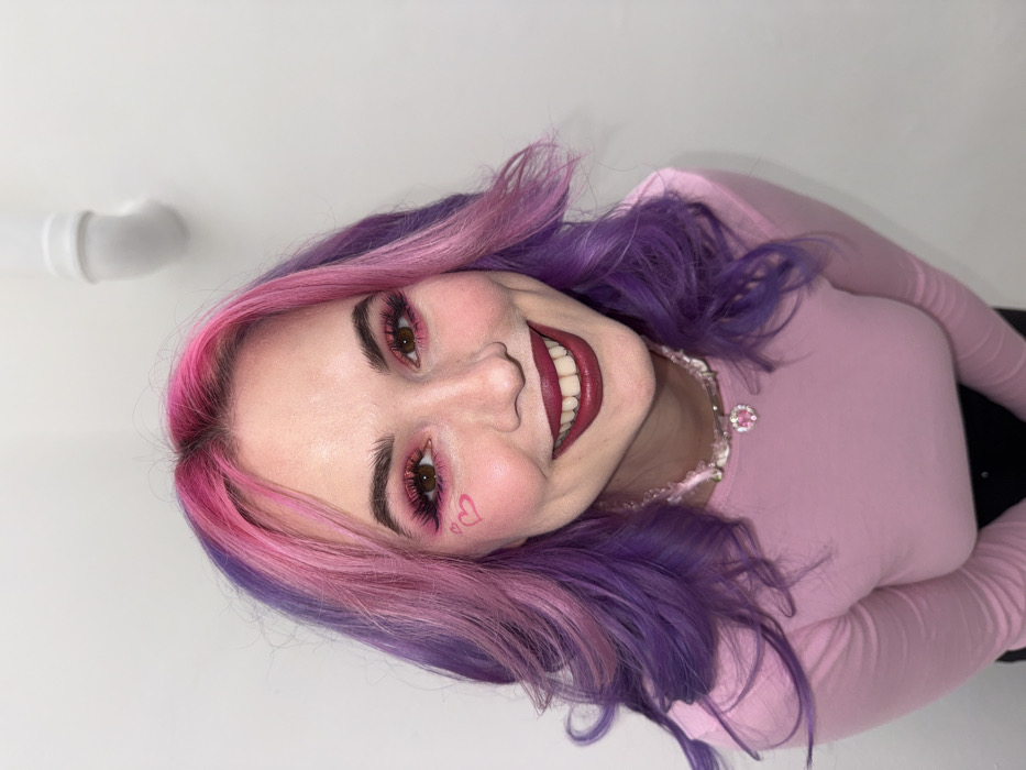 A female subject, appearing to be a young adult, is depicted from the upper chest upward, smiling directly at the camera. Her hair is styled in waves, with the roots and front sections dyed vibrant pink, transitioning to purple towards the sides and ends. She has prominent eye makeup, including pink eyeshadow and dark eyeliner, and a small pink heart is drawn on her left cheekbone. Her lips are covered in dark red lipstick, and she displays a broad smile revealing her upper teeth. She wears a light pink long-sleeved top and a choker-style necklace featuring clear beads and a central pink heart-shaped pendant. The background is a plain, light-colored wall. In the upper right quadrant of the image, a white, exposed plumbing pipe or conduit is visible, bent at a 90-degree angle. No specific identifying markers for the City of Zagreb, Croatia, are present within the visual frame.