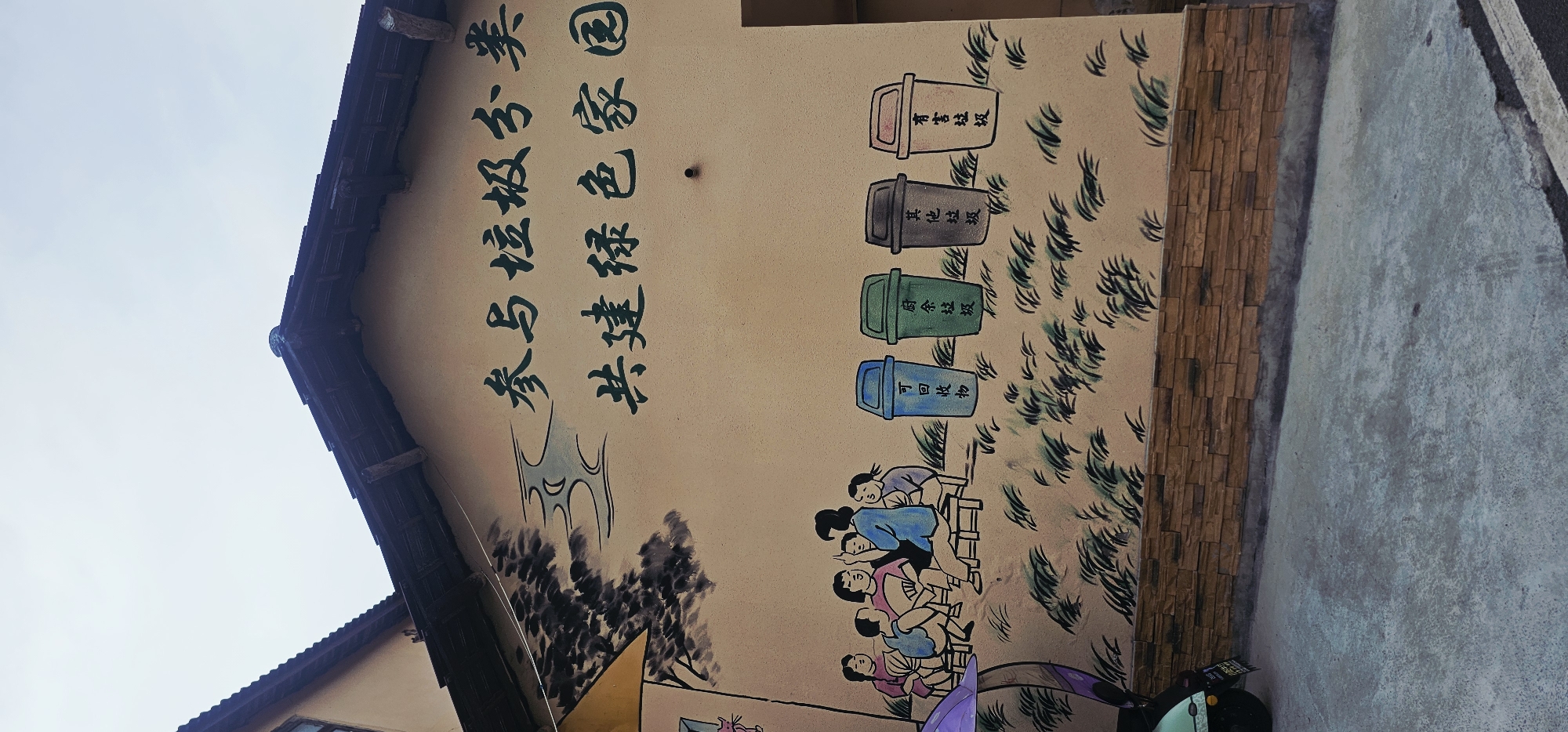 The image shows a mural on the exterior wall of a building in Shapingba District, China. The mural depicts a group of people sitting on benches and talking, with several colorful trash cans lined up in front of them. The trash cans are labeled with Chinese characters indicating different types of waste: "recyclable waste," "kitchen waste," "other waste," and "hazardous waste." Above the mural, the wall is painted with large Chinese characters that translate to "Participate in garbage classification and build a green home together." The building has a tiled roof and a brick base, and the ground in front of it is paved with concrete. The scene appears to be outdoors, with a cloudy sky overhead.