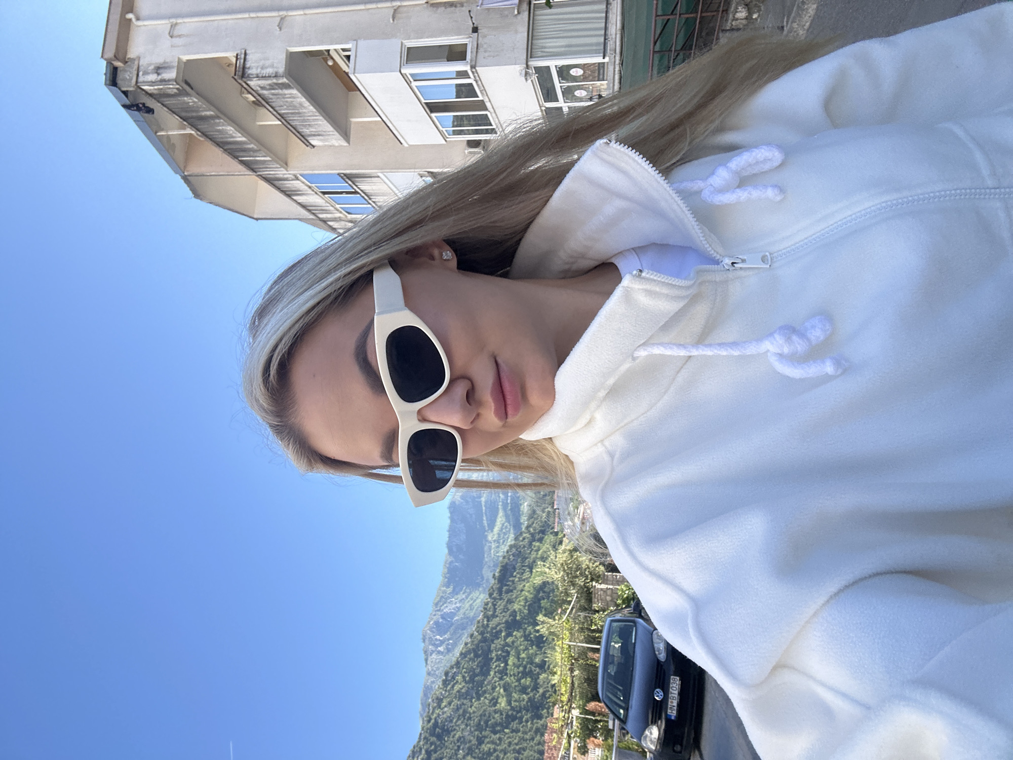 A sideways portrait of a woman wearing a white fleece jacket and white-rimmed sunglasses is captured against a backdrop of blue sky, a building, and a mountain range. The woman is positioned in the foreground, with her face turned towards the camera. Her blonde hair is partially visible behind her sunglasses. The white fleece jacket she wears has a zippered front and drawstrings at the collar.
Behind her, a light-colored building with windows is visible in the upper left portion of the frame. In the lower right corner, a black car is parked on the side of a road, and a mountain range covered in green vegetation is seen in the distance. The location is likely near Budva, Montenegro, given the mountainous backdrop and urban elements.
