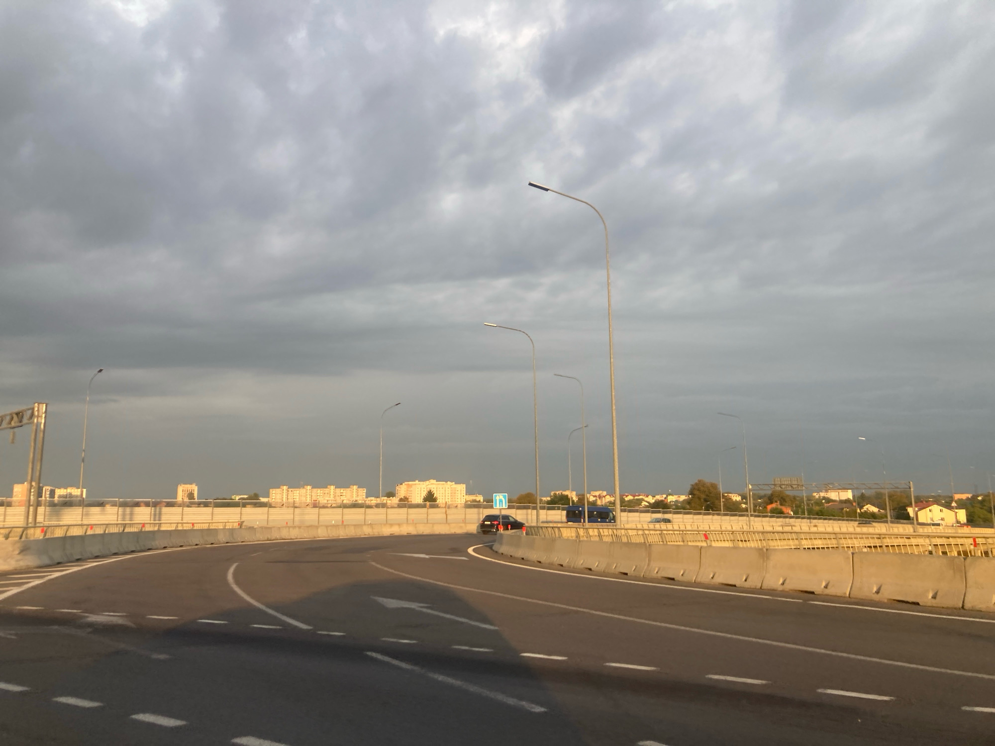 The image depicts a highway overpass in Brest, Belarus. The sky is overcast with gray clouds. On the highway, there are several vehicles including a dark car and a blue van. The highway is lined with concrete barriers and streetlights. In the background, there are apartment buildings and houses, suggesting an urban or suburban setting. The sun is low in the sky, casting a warm light on some parts of the scene, particularly the buildings in the distance.