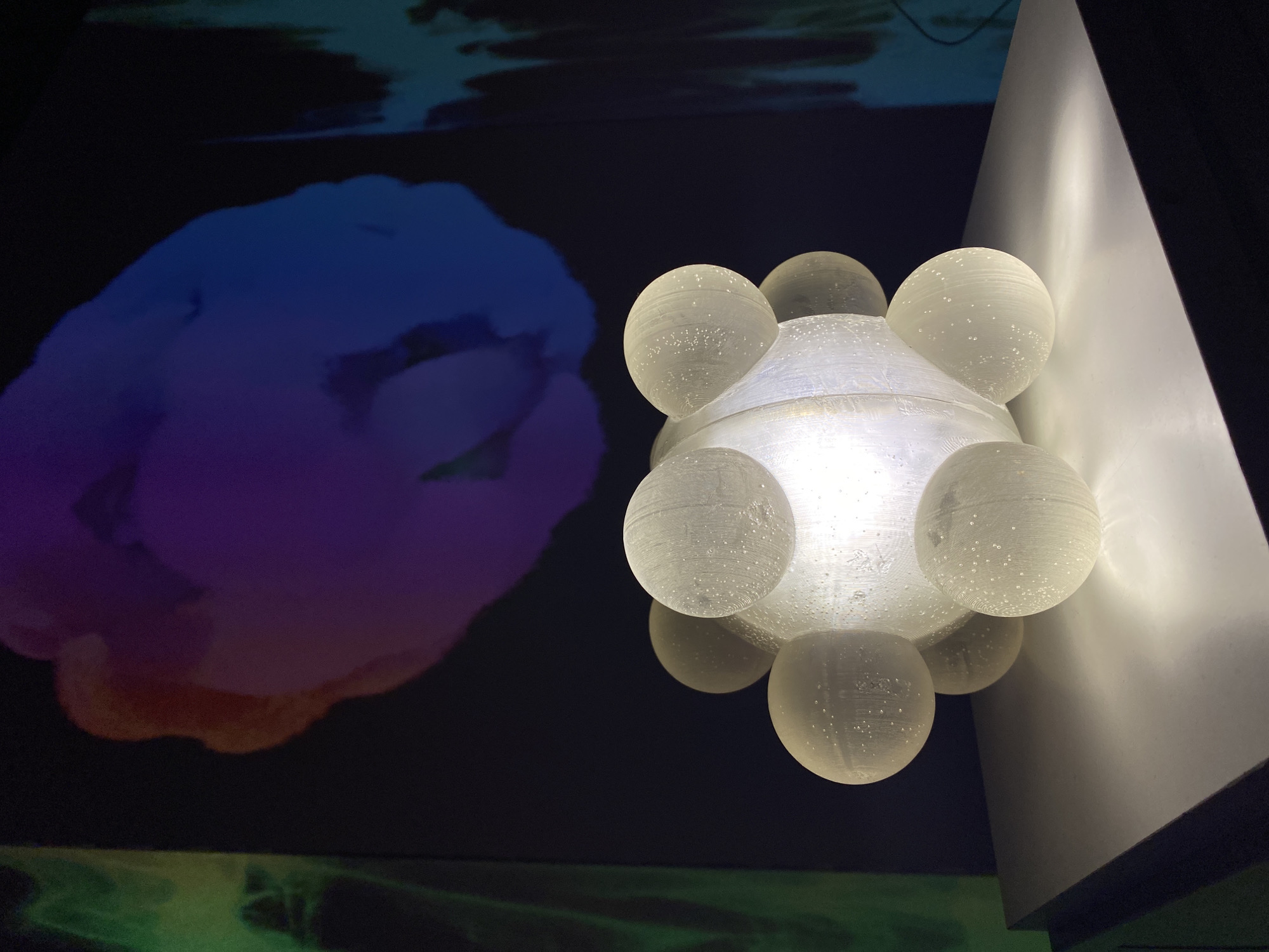 The image captures an art installation or exhibit, with a unique lighting fixture as the focal point. The location is Yekaterinburg, Russia.
The lighting fixture is composed of a central structure, possibly a translucent or opaque dome, surrounded by multiple spherical bulbs or globes. The spheres are arranged in a cluster, creating a complex, geometric form. The fixture is illuminated from within, casting a bright, warm light that highlights the textures and details of the material.
In the background, there is a large projection or display. The image shows a cloud-like form with gradients of blue, purple, and orange. The lower part of the background has a green tint.
The scene is set in what appears to be an indoor space. The lighting is subdued, with the main source of illumination coming from the central fixture. There is also light reflecting off a white, angled surface to the right.