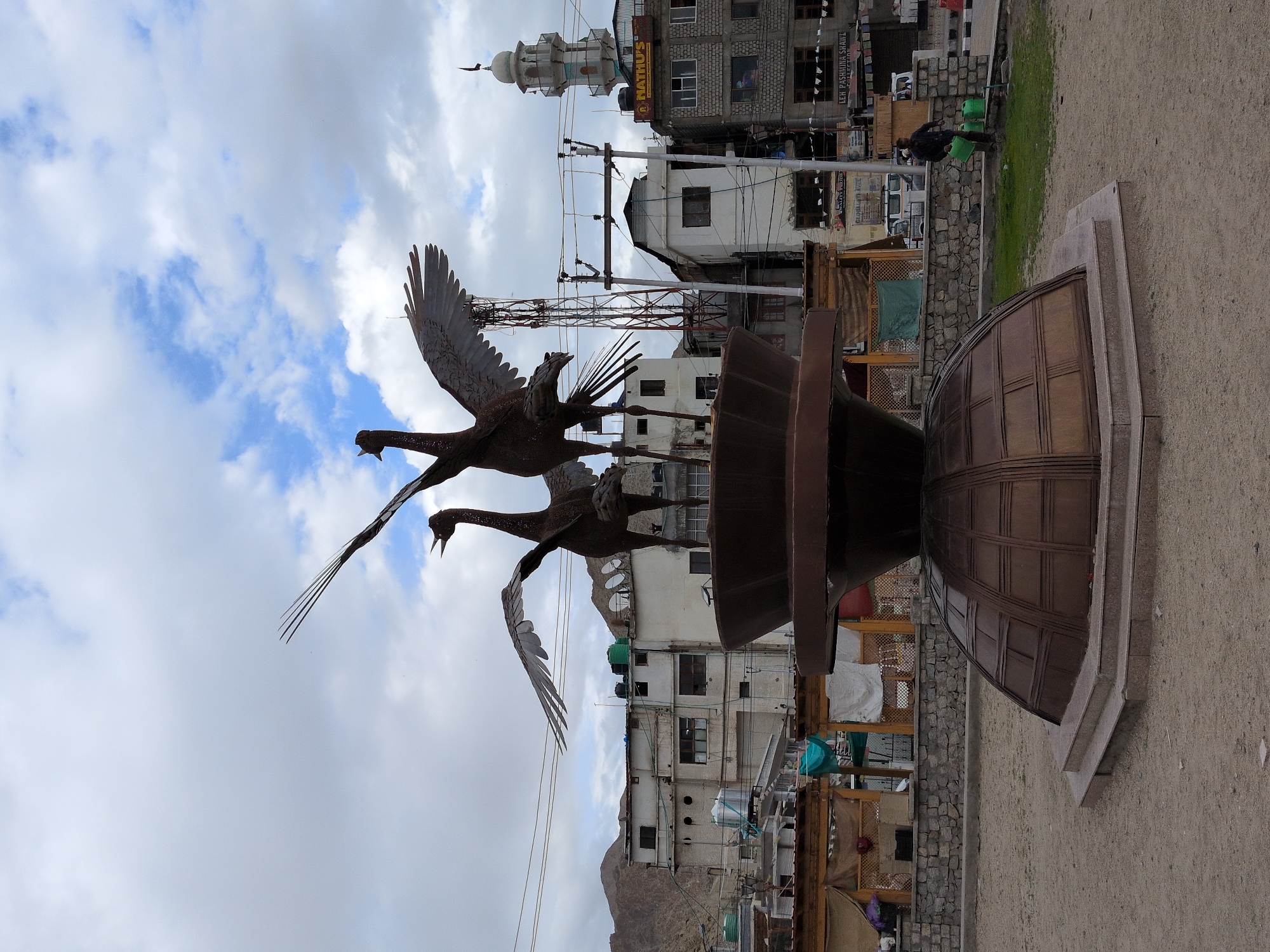 In Leh, India, a bronze sculpture of two cranes with outstretched wings stands in a dusty plaza. The sculpture is mounted on a multi-tiered, dome-shaped pedestal. Behind the sculpture, a street is lined with buildings, including structures with flat roofs and a mosque with a minaret. A telecommunications tower is also visible. The sky is cloudy, with patches of blue. A person is watering a patch of grass to the right of the sculpture, holding two green watering cans. The ground is covered in sand and gravel.