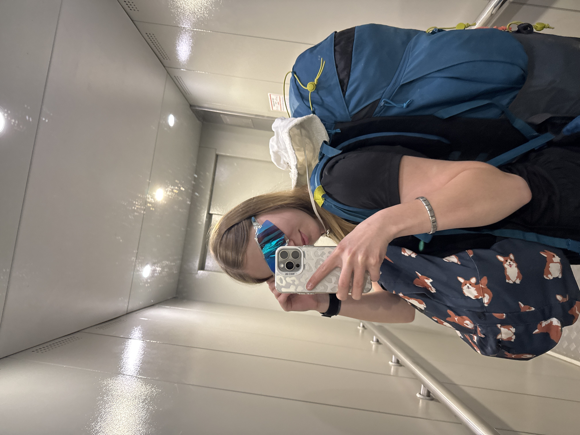 A person is holding a smartphone and taking a selfie. They are wearing a dark t-shirt, patterned pants with Corgis, a watch, and a silver bracelet. They are also wearing large, reflective sunglasses and carrying a blue and black backpack. The person is inside a small, enclosed space with white walls and a metallic floor. The location is most likely not in the city of Chaikovsky, Russia based on the image content and setting.