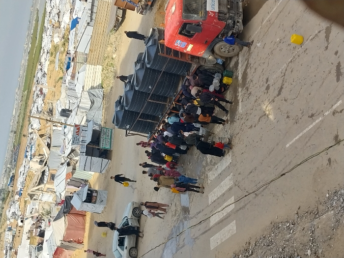 The image depicts an outdoor scene in a temporary settlement within Khan Yunis, Palestinian Territory. A red flatbed truck, visibly branded with "UNICEF" on its front fender, is parked on a dirt road, laden with at least eight large, black cylindrical water tanks. A crowd of approximately 30-40 individuals, consisting of adults and children, surrounds the rear and right side of the truck, actively collecting water into various containers, including yellow jerrycans, white buckets, and other plastic vessels. The ground immediately around the truck is wet and muddy from water spillage.
In the mid-ground, a light-colored sedan is parked on the left side of the road with its front and rear left doors open; two individuals are visible near it, one holding a yellow jerrycan and walking away. The background features an extensive, densely packed area of makeshift shelters and tents constructed from tarpaulins, fabric, and assorted debris, stretching into the distance under an overcast sky. Several small, temporary structures resembling stalls or shops are present among the tents, one displaying Arabic text that includes "المتحدة" (United). A faded white zebra crossing is marked on the paved section of the road in the foreground, adjacent to an unpaved shoulder. Green fields are faintly discernible on the distant horizon beyond the settlement.