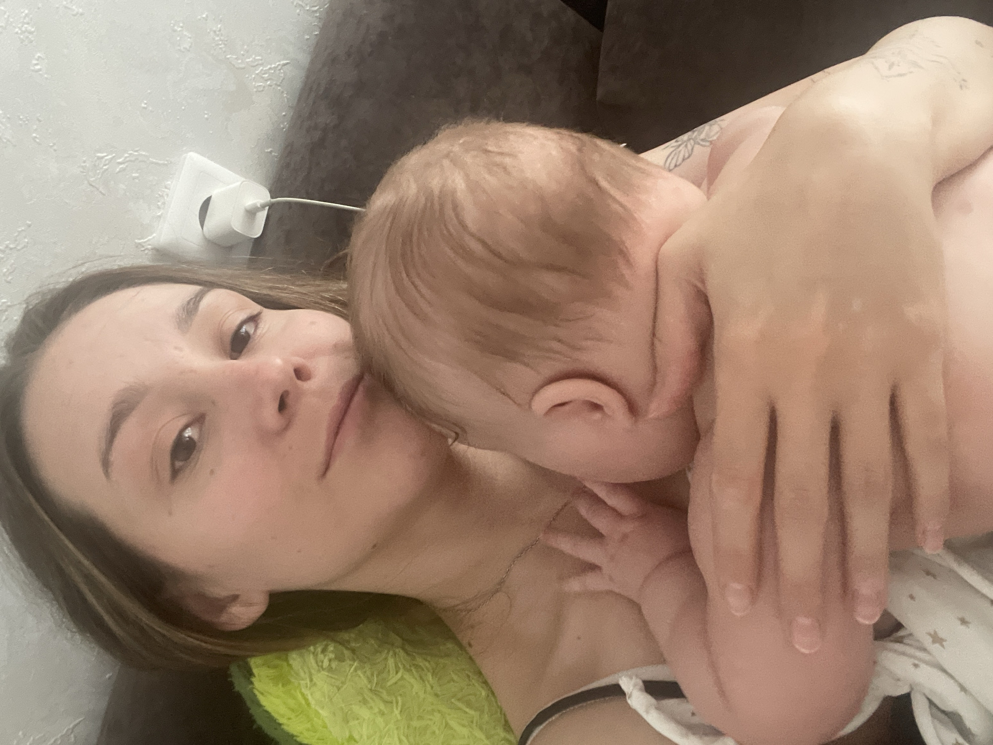 The image shows a woman and a baby lying down, possibly on a bed or couch. The woman is looking at the camera with a calm expression. The baby is nuzzling into the woman's neck, with its face partially obscured. The baby's hand is resting on the woman's chest. The background is blurred, but suggests an indoor setting with a textured wall and a grey couch. A white electrical outlet with a charger plugged in is visible on the wall. The lighting appears to be indoor lighting, suggesting it could be any time of day. The woman has a tattoo on her shoulder. The overall mood of the image is intimate and peaceful, capturing a tender moment between mother and child. No text is visible in the image.
