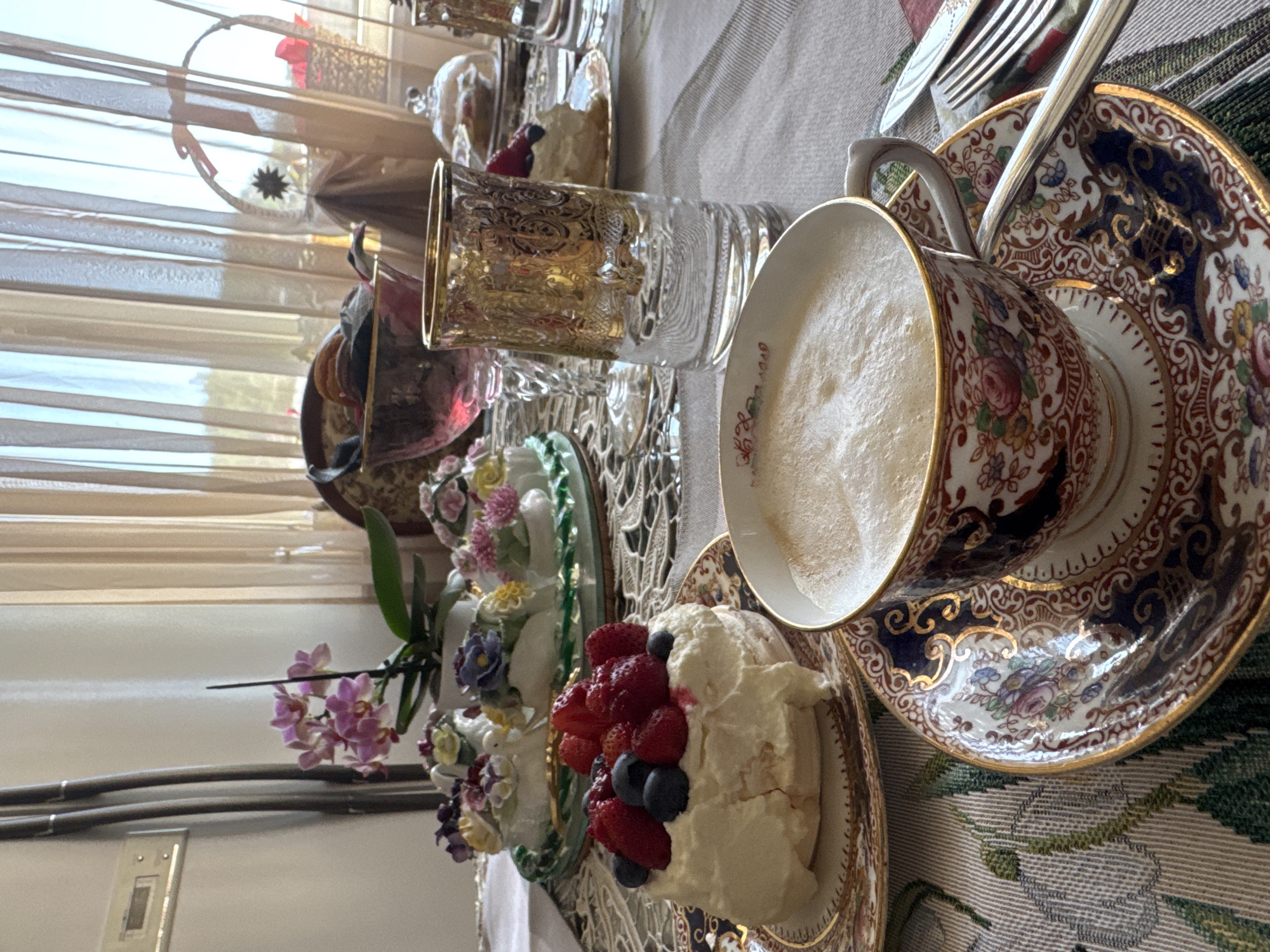 A formal afternoon tea setup is depicted on a table with a patterned tablecloth. In the foreground, a delicate porcelain teacup and saucer, adorned with intricate floral designs and gold trim, hold a frothy cappuccino. Beside it, a silver spoon rests on the saucer. To the left, a small pavlova topped with fresh strawberries and blueberries sits on a matching ornate plate. In the background, a white porcelain dish holds miniature floral arrangements, and a clear glass with gold embellishments stands next to a pink-stemmed glass. Beyond the table, sheer beige curtains are drawn, revealing a sunlit outdoor scene. The overall atmosphere suggests a refined and elegant setting, possibly a cafe or a home dining room, where one might enjoy a leisurely tea. There is no indication of the specific location in Plovdiv, Bulgaria.