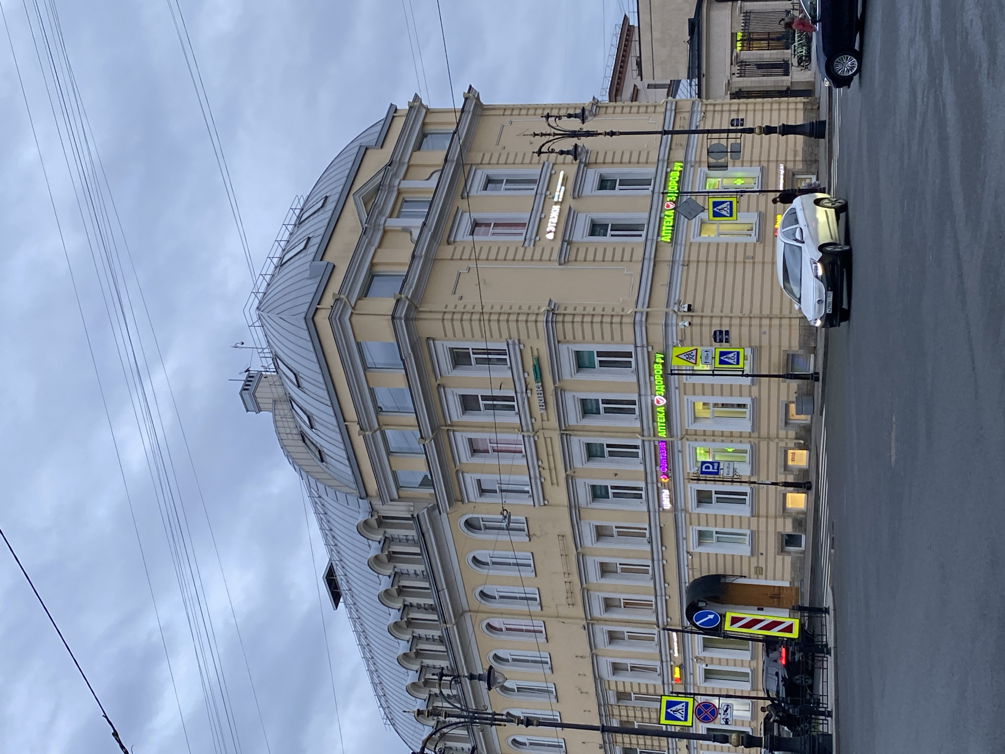 The image depicts a street scene in Saint Petersburg, Russia, under an overcast sky. A large, multi-story building with a curved roofline dominates the frame. The building has a yellow facade with white window frames. Several signs, including neon green signs that read "Аптека Здоров.ру" (Pharmacy Health.ru), are affixed to the building's exterior. A white car is in motion on the street in front of the building. There are street lamps and pedestrian crossing signs along the sidewalk. Power lines are visible in the upper left of the frame. A black car is parked further down the street. Pedestrians can be seen near the parked car.