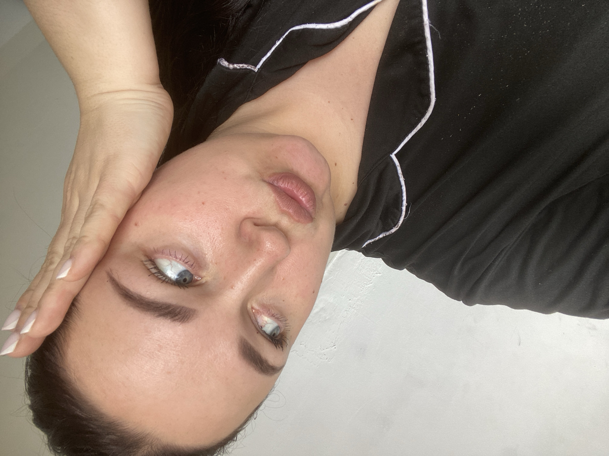 The media file presents an indoor portrait of a woman, captured from an overhead perspective, possibly in Sochi, Russia. The subject, a woman with fair skin, dark hair, and blue eyes, is lying down, appearing to be on her back with her head slightly tilted. Her right hand rests near her forehead or temple, revealing long, light-colored fingernails with a distinct pointed tip, likely a French manicure. Her expression is neutral, with her eyes looking upwards and to the side, away from the direct camera lens, conveying a contemplative or relaxed mood.
She is dressed in a black top, possibly pajamas, featuring prominent white piping along the neckline. Close-up examinations of the garment reveal small white fibers or lint scattered across the black fabric. Details of her appearance include natural pinkish-brown lips, dark and well-defined eyebrows, and visible eyelashes on her blue eyes. Her skin shows fine lines on the inner wrist and small moles or freckles on her neck and chest area. The background is a plain, light-colored surface, appearing to be a wall or bedsheet, with a faint gray mark visible in one of the crop views. The lighting is soft and even, typical of an indoor setting with diffused light, but no specific time of day is indicated. There is no visible text in the image.