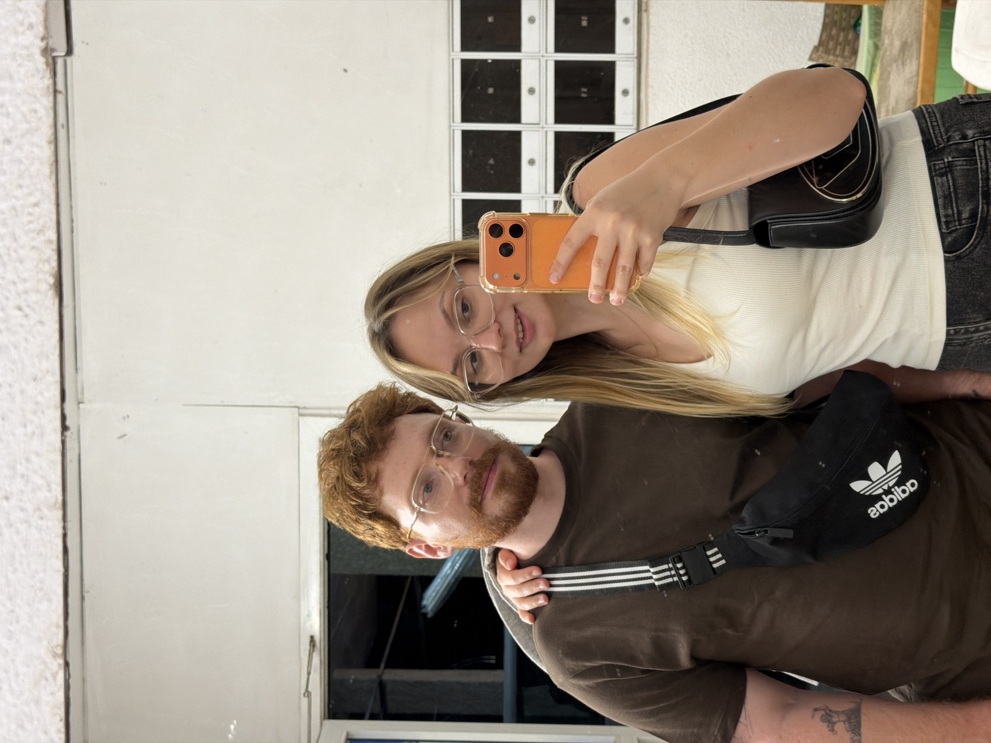 A young couple is reflected in a mirror, likely taking a selfie. The woman, with blonde hair and glasses, holds an orange phone. She wears a white top and a black shoulder bag. The man, with red hair and a beard, wears glasses and a brown t-shirt, with a black Adidas fanny pack across his chest. A tattoo is visible on his arm. Behind them is a white wall with square black and white panels. The reflection also shows part of a window and greenery. The location appears to be indoors. This is in Rehovot, Israel, given the prompt.