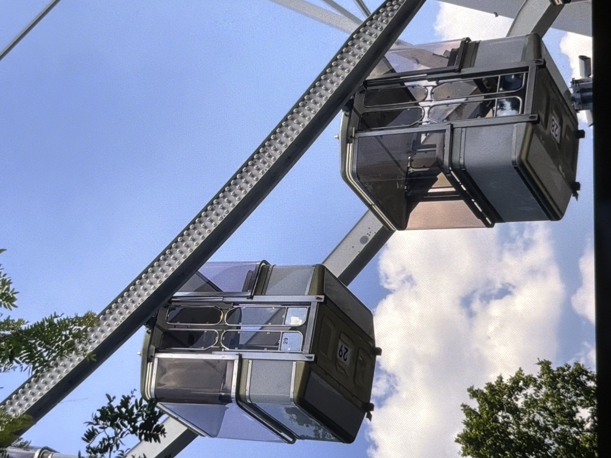 The image shows two cabins of a Ferris wheel against a blue sky with some white clouds. The cabins are made of metal and glass and appear to be empty. One cabin has the number "29" visible on its side, and the other has the number "82". The Ferris wheel structure is made of metal and has a series of studs along its beam. Green trees are visible at the bottom of the frame, suggesting an outdoor setting, likely a park or amusement area. The lighting indicates it is daytime.
