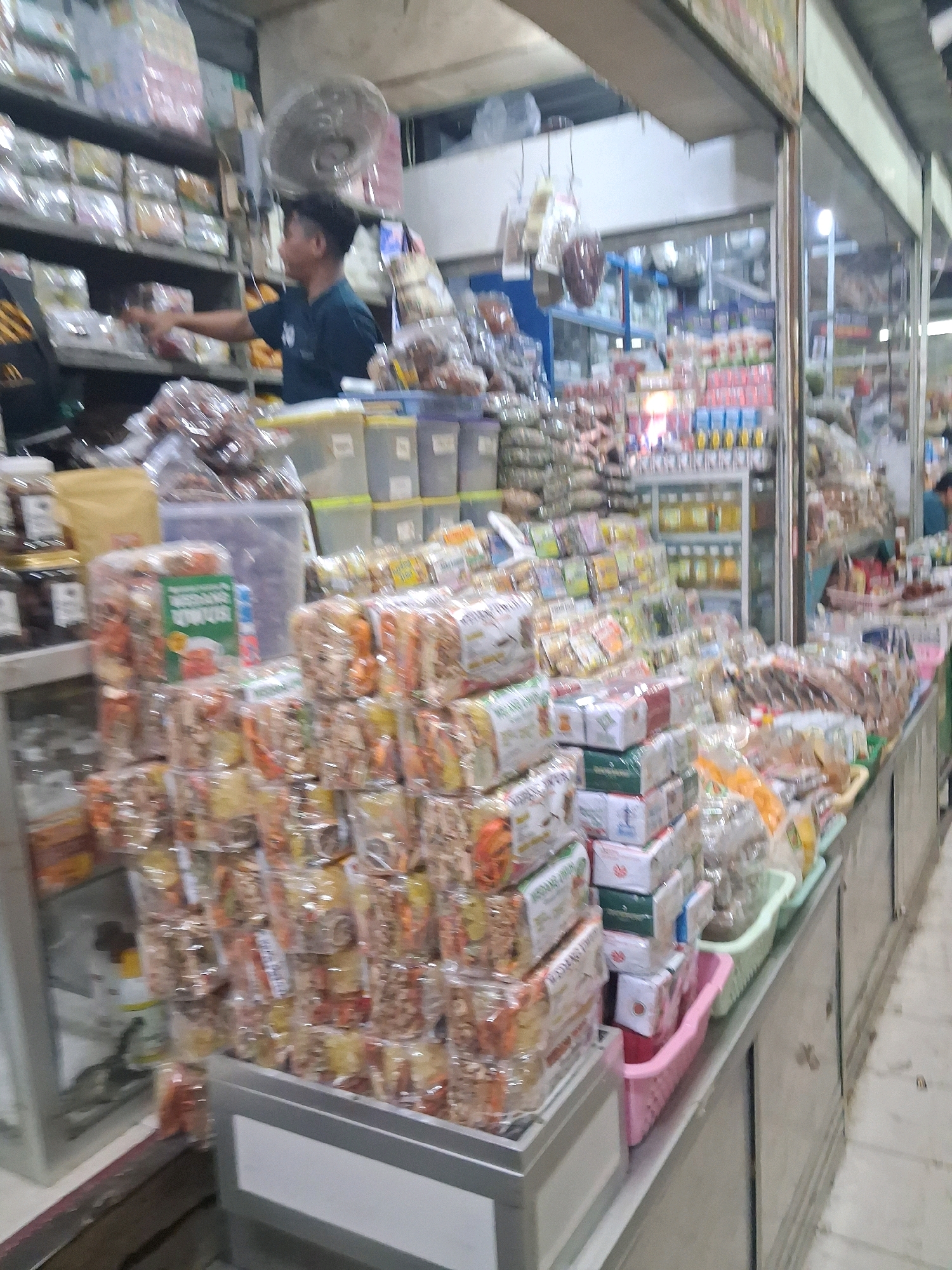 A man is standing behind a stall in a market in Surakarta, Indonesia. The stall is filled with various food items, mostly packaged snacks. The man, wearing a dark blue t-shirt, is reaching out to a shelf with his left hand. The stall has multiple shelves stacked with products in clear plastic packaging and some boxes. In the background, there are more shelves, a fan on the wall, and other stalls. The overall impression is a busy marketplace scene.