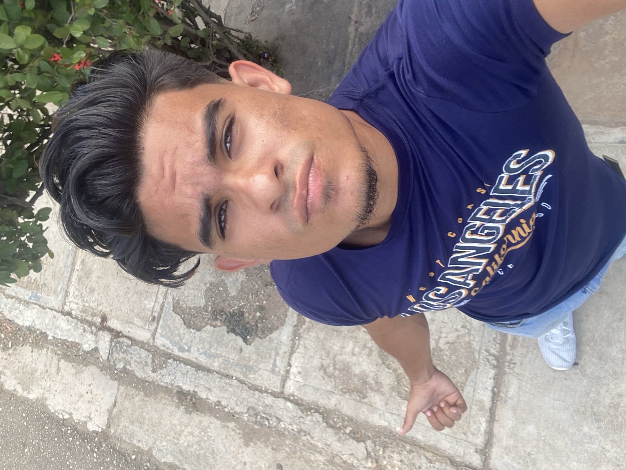 A dark-haired man is shown in an upward-facing selfie, his shirt bearing the words "Los Angeles California" in decorative script. The man is light-skinned with dark hair and a short beard. He stands on a stone walkway in an outdoor environment with a few green leafy plants near the top left corner. The location is identified as Juan Griego, Venezuela. He is wearing light blue jeans and sneakers. He is extending his hand toward the camera, making a thumbs-down gesture.