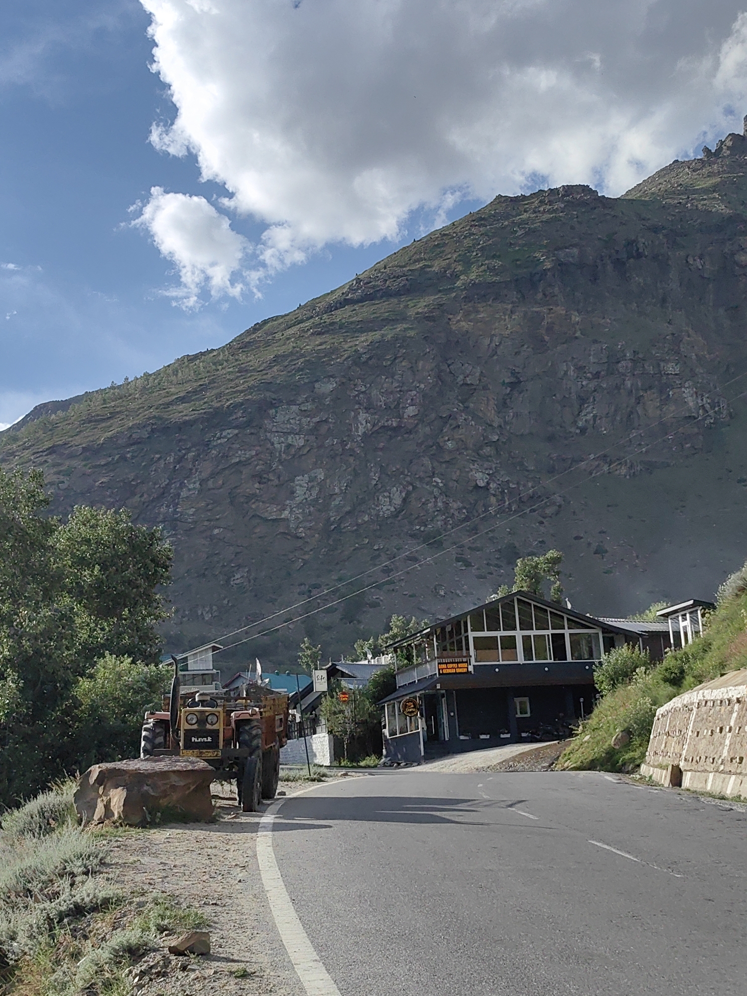 A tractor is parked on the side of a paved road in a mountainous area, possibly Yamling, India. The tractor is orange and yellow and is loaded with cargo in its rear trailer. A large rock is placed in front of the tractor's wheels. The road curves to the right. In the background, there are several buildings with dark siding and large windows. A steep, rocky mountain covered in sparse vegetation rises behind the buildings. The sky is blue with scattered white clouds. The overall scene suggests a rural or remote location with natural beauty.
