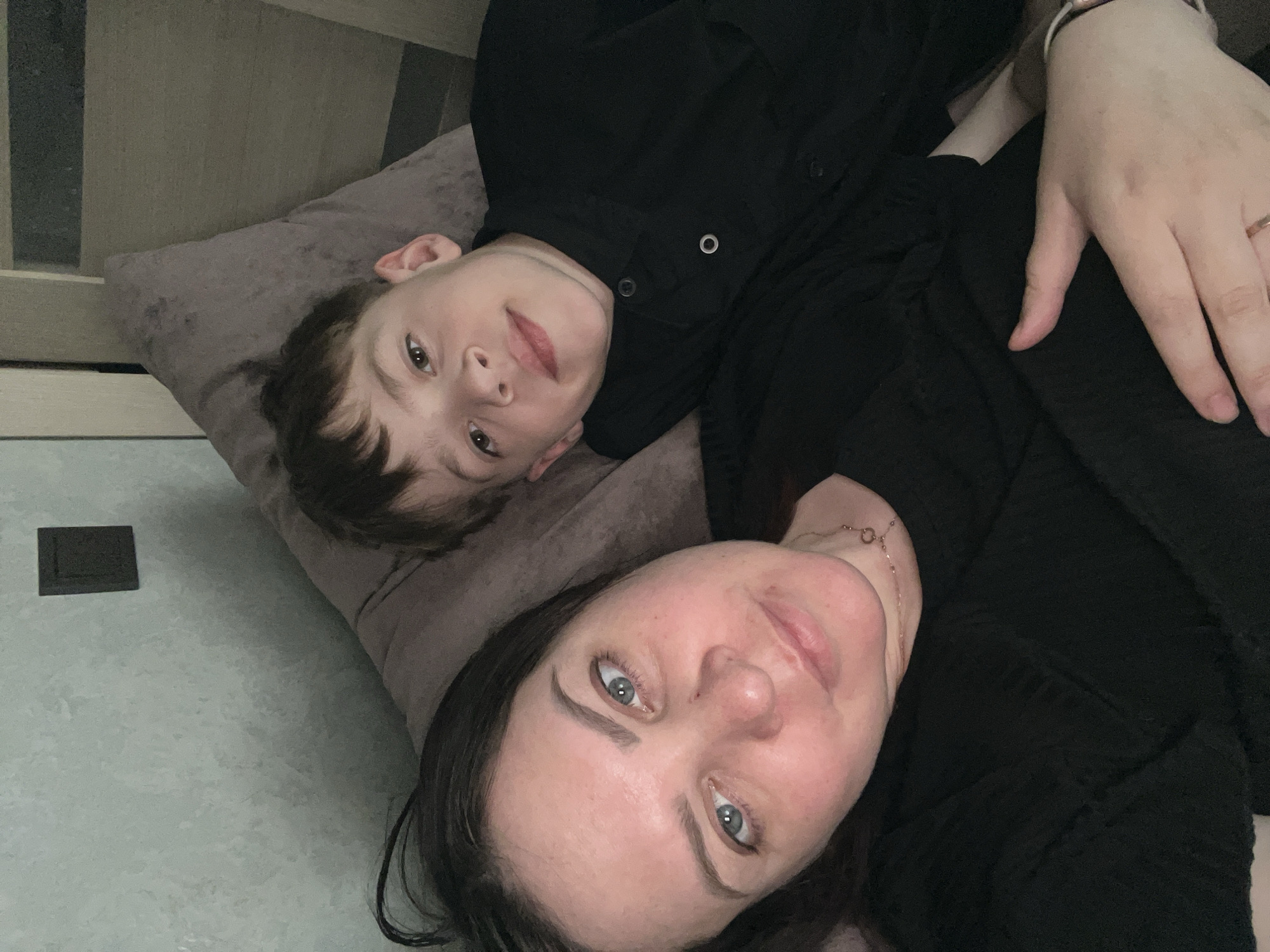 A photograph shows a woman and a boy lying down. The boy rests on a pillow, and his face is visible. He wears a black button-up shirt. The woman is next to him, also lying down. She wears black clothing. A hand wearing a watch and a ring is visible. The background includes a tiled floor and a section of furniture. The location information, Noviy, Russia, is given separately.