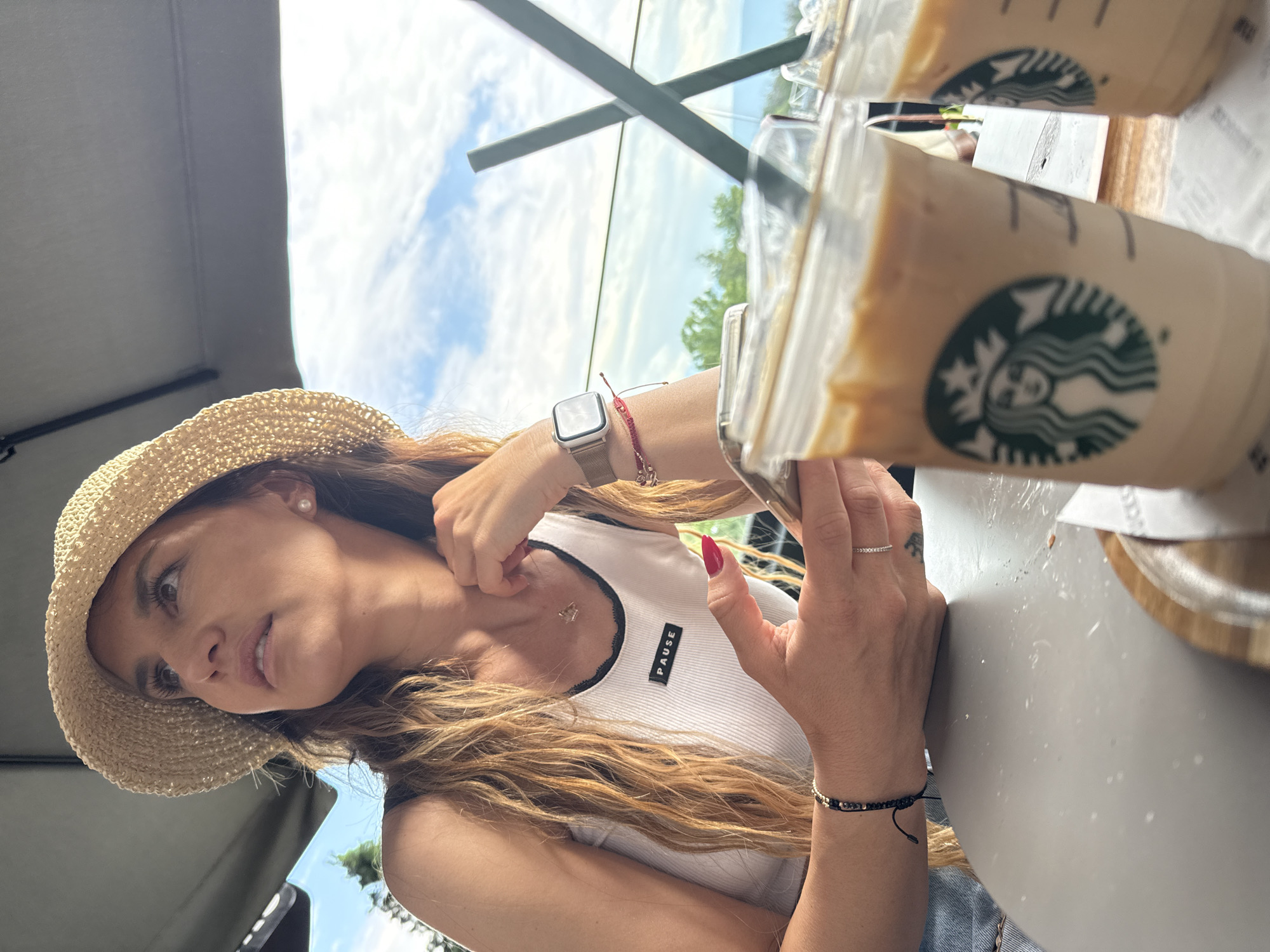 An adult female is seated outdoors, partially shaded by a grey umbrella, under a blue sky with scattered white clouds, in an elevated position consistent with a viewpoint in Plovdiv, Bulgaria. She has long, wavy light brown hair and is wearing a straw hat, a white ribbed tank top with black trim and a black rectangular "PAUSE" patch, a delicate necklace, an Apple Watch with a metallic mesh band, a red string bracelet, and a black beaded bracelet on her left wrist. On her left hand, she wears a ring with small stones on her ring finger and has a small tattoo on the side of the same finger. Her fingernails are painted red. She holds a smartphone in her left hand and looks upwards and to the right, with a contemplative expression. On the grey round table in front of her are two clear plastic Starbucks cups with green straws, containing iced coffee, resting on a wooden coaster. In the background, beyond a glass railing, is a panoramic view of distant green hills and trees under the cloudy sky.