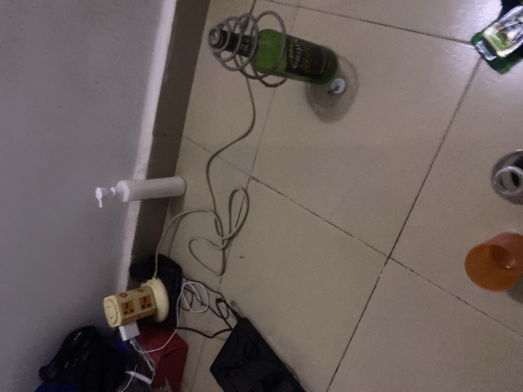 In a room with tiled flooring and a beige wall, a green beer bottle is secured within a wire cage attached to a cord, positioned on the floor. Nearby, a white dispenser bottle stands on the floor next to the wall. Electrical wires snake across the floor, connecting to a yellow power strip with multiple outlets and a red device. A black bag and a black portable device are also visible. In the foreground, an orange cup and a silver-colored object are present. The scene is likely located in Howayi, Nigeria, a residential area.