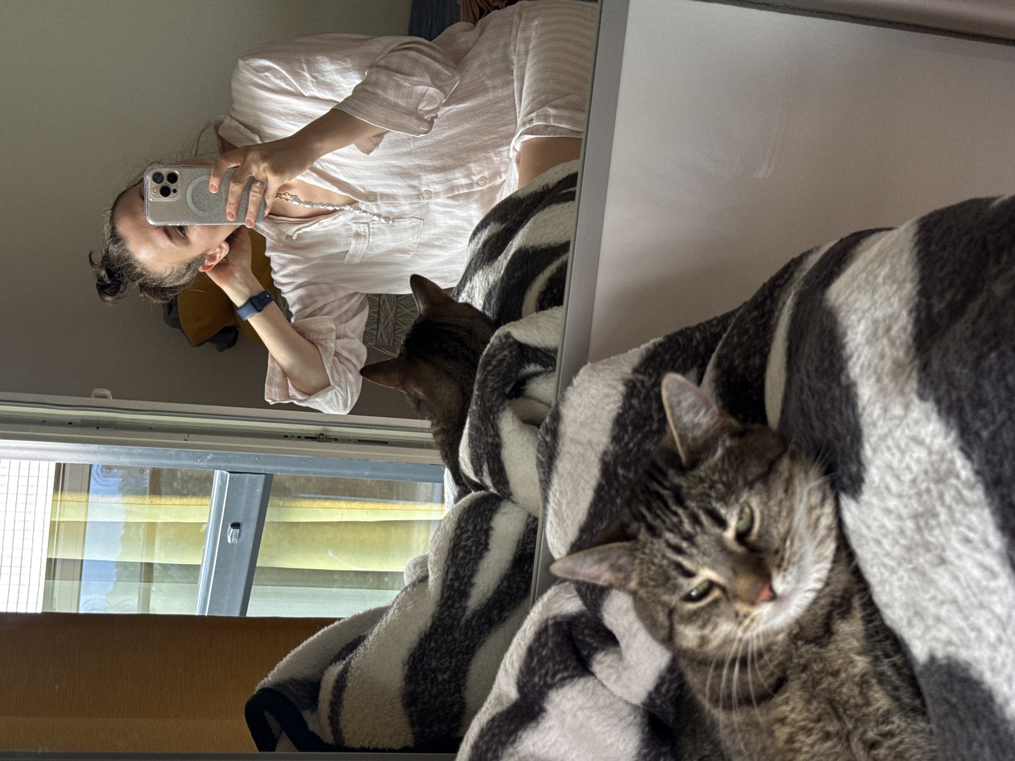 In Saint Petersburg, Russia, a woman is captured in a mirror selfie, holding a smartphone in her right hand and adjusting her hair with her left. She is wearing a light pink striped pajama set and a dark blue fitness tracker on her left wrist. A pearl necklace hangs from her neck. To her left, a tabby cat with green eyes is resting on a black and white striped blanket. Another cat, with a dark brown back, is partially visible behind the woman, also on a blanket. The scene appears to be indoors, possibly a bedroom, with a window and blinds visible in the background. The reflection in the mirror shows the woman and cats, along with a yellow and green striped curtain. The overall impression is a relaxed, domestic moment.