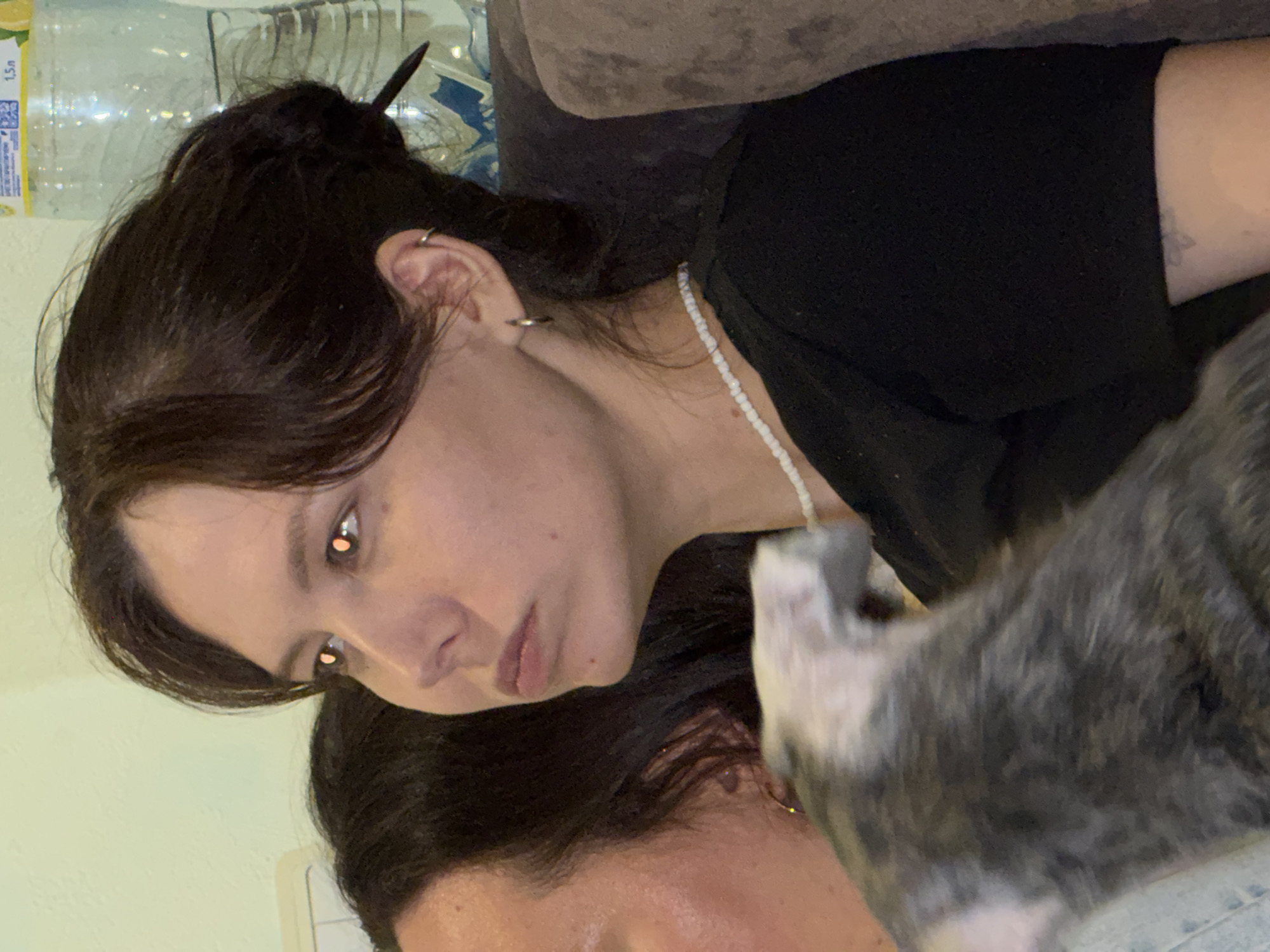 A woman with dark hair pulled back, wearing a black shirt and a white beaded necklace, is looking to her left. A grey and white cat is partially visible in the foreground, near her neck. A bottle of water is visible in the background. The location is unidentifiable as Chaikovsky, Russia.