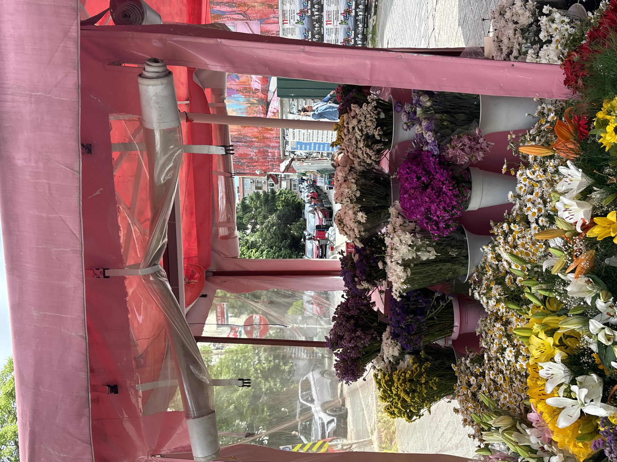 A vibrant flower display occupies the right side of the frame, featuring diverse blooms of white, yellow, orange, and purple hues arranged in white buckets. To the left, a pink canopy structure with a transparent plastic covering partially obscures the view of a street. In the background, the scene reveals a street with cars and buildings. This image was taken in the city of Kadıköy, Turkey.