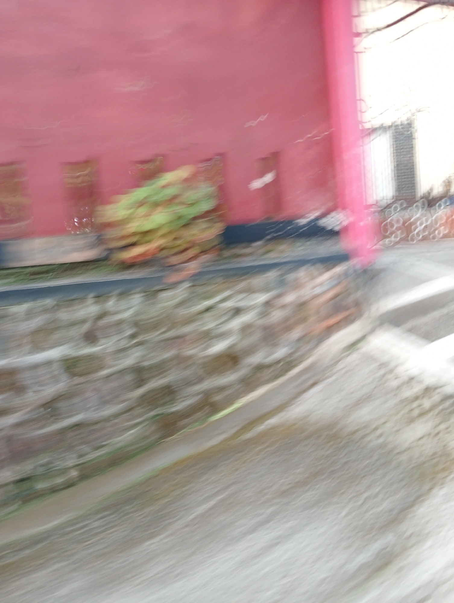 The image depicts a blurry outdoor scene with a red building in the background. In the middle ground, a textured stone wall is visible. The foreground shows a sloped, light-colored surface, possibly a path or pavement. There are hints of greenery and architectural elements, but due to the motion blur, specific details are indistinct. The location is identified as Krajan Kidul, Indonesia.