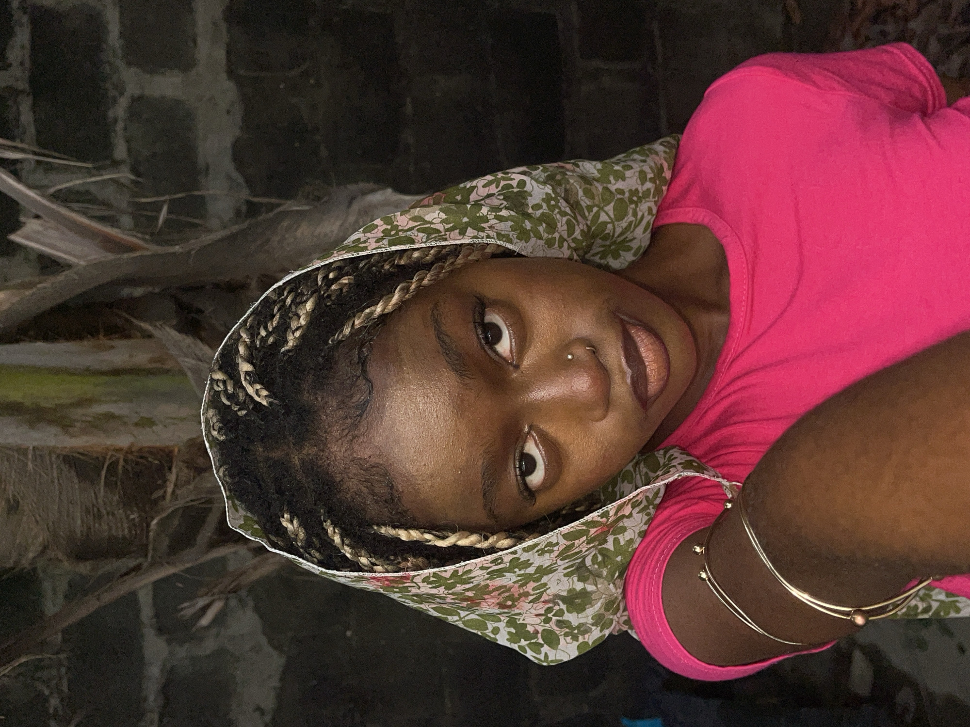 An eye-level portrait features a person, likely female, with dark skin, positioned at a 90-degree angle to the right, looking directly at the viewer. The subject wears a vibrant hot pink t-shirt and a light-colored head covering with a green and pink floral or leafy pattern. Their hair is styled in dark braids with lighter, possibly blonde, sections interwoven. Notable facial features include wide, dark eyes, dark eyebrows, and full lips with a subtle, possibly lined, light brown or nude lip color. A small silver nose piercing is visible on the left nostril. On the right arm, two thin gold-colored bangles or bracelets are worn.
The setting is dark, suggesting evening or nighttime conditions, with the subject well-lit in contrast to the blurry, dimly lit background. Behind the person, a textured wall or structure, possibly made of concrete blocks or stone, is visible. Below that, indistinct dark shapes resembling branches or debris are present. The overall atmosphere is intimate, given the direct gaze and close-up nature of the shot. The expression is calm and engaging. No visible text is present. The context provided indicates the location is Ado, Nigeria.