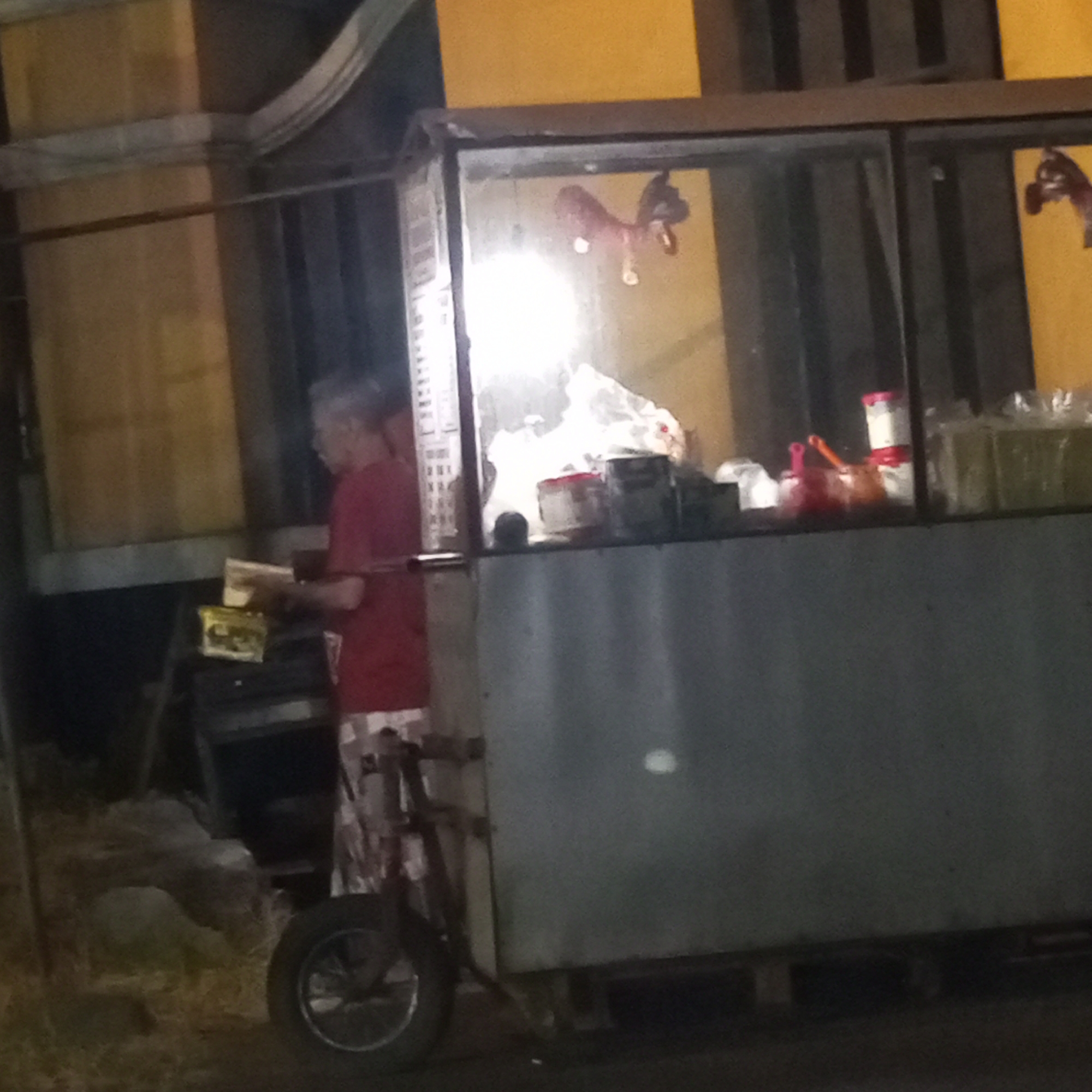 The image depicts a street vendor stall at night in Sanggrahan, Indonesia. A person in a red shirt and patterned shorts is standing behind a food cart, appearing to handle items. The cart is a metal structure with glass windows, displaying various goods including bottled beverages. The background shows a yellow wall and some interior structures. The overall scene is dimly lit, suggesting it is nighttime.