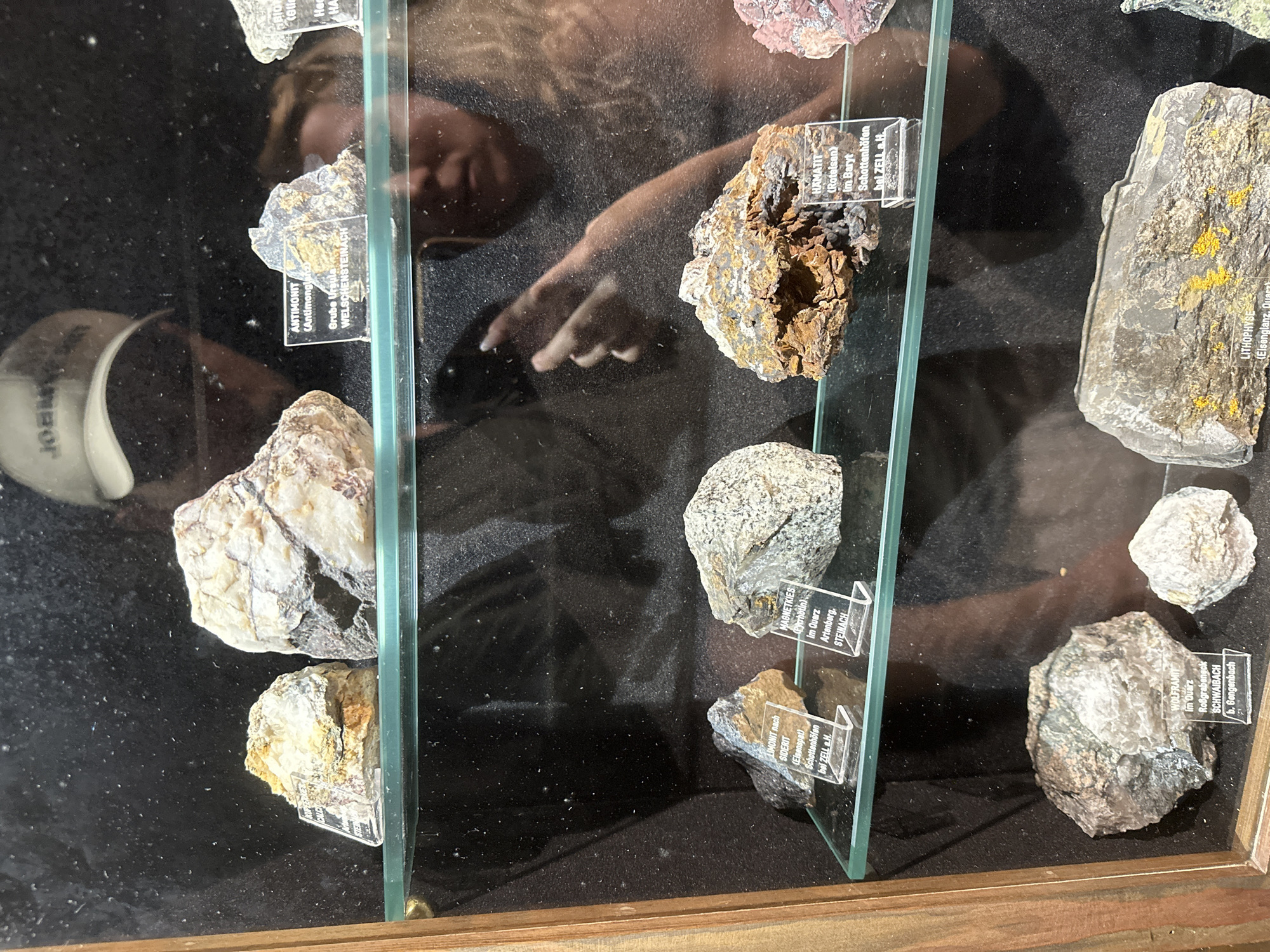 This media file shows a display case containing various mineral samples, likely in a museum or a collector's exhibit in Schonach im Schwarzwald, Germany. The display case has a wooden frame and glass shelves, with several minerals arranged neatly on a dark background. Each mineral specimen has a small label in front of it, identifying the mineral and its origin.
Visible text on the labels includes: "ANTIMONIT (Antimong Grube Ursula WELSCHENSTEINACH", "LIMONIT nach SIDERIT (Eisenspat) Schottenhöfen bel ZELL a.H.", "HAMATIT (Rotelsen) Im Baryt Schottenhöfen bel ZELL a.H.", "MAGNE TKIESS (Pyrrhotin) Im Quarz Artenberg. STEINACH", "WOLFRAMIT Im Quarz Roßgrabeneck SCHWAIBACH b. Gengenbach", and "LITHOPHYSE (Elsenglanz, Quarz)". The reflection on the glass reveals a person, likely a visitor, looking at the exhibit. The person's hands are visible, suggesting they might be pointing at a specimen or interacting with the display in some way. The lighting suggests an indoor setting, possibly during the day. The overall scene conveys an educational or recreational context related to geology and mineralogy.