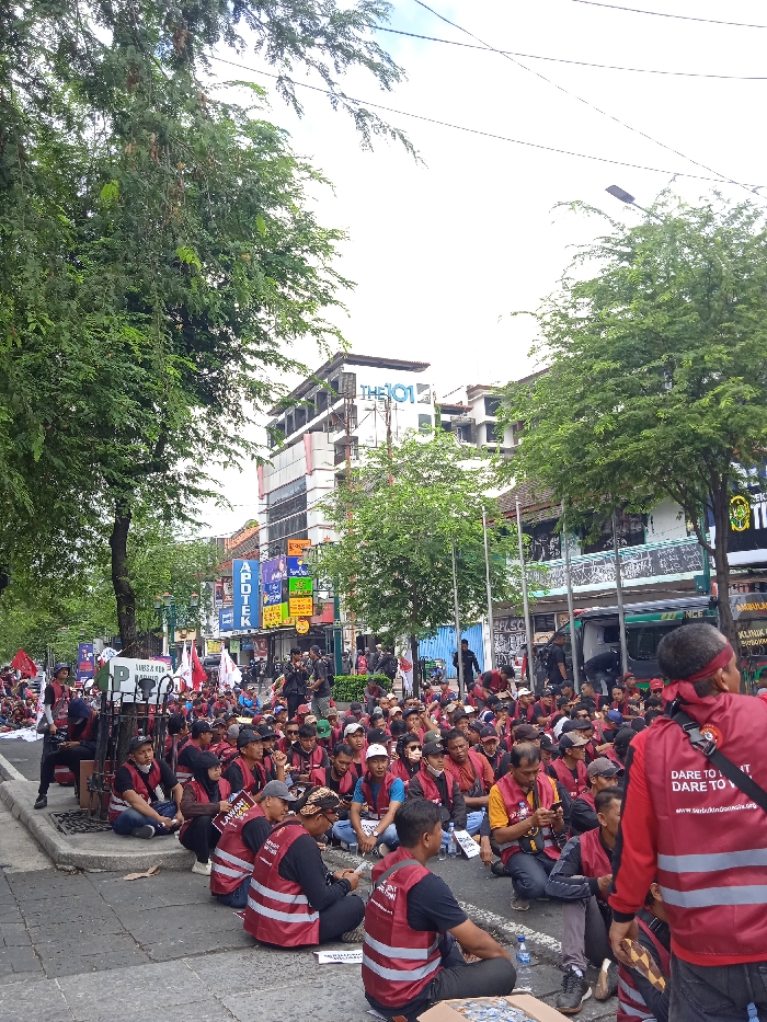 The image shows a street scene in Yogyakarta, Indonesia, taken at the coordinates -7.784 latitude and 110.367 longitude. A group of people, mostly men, are seated on the sidewalk in front of a building, many wearing red vests with the words "Dare to Fight for Justice". They are all facing the camera, some holding signs, banners, and flags. Several individuals are standing behind the seated group, observing the scene. The buildings in the background appear to be commercial, with signage and windows. There are trees on either side of the street, and power lines are visible above. The overall scene suggests a gathering of people engaged in a peaceful protest or demonstration.