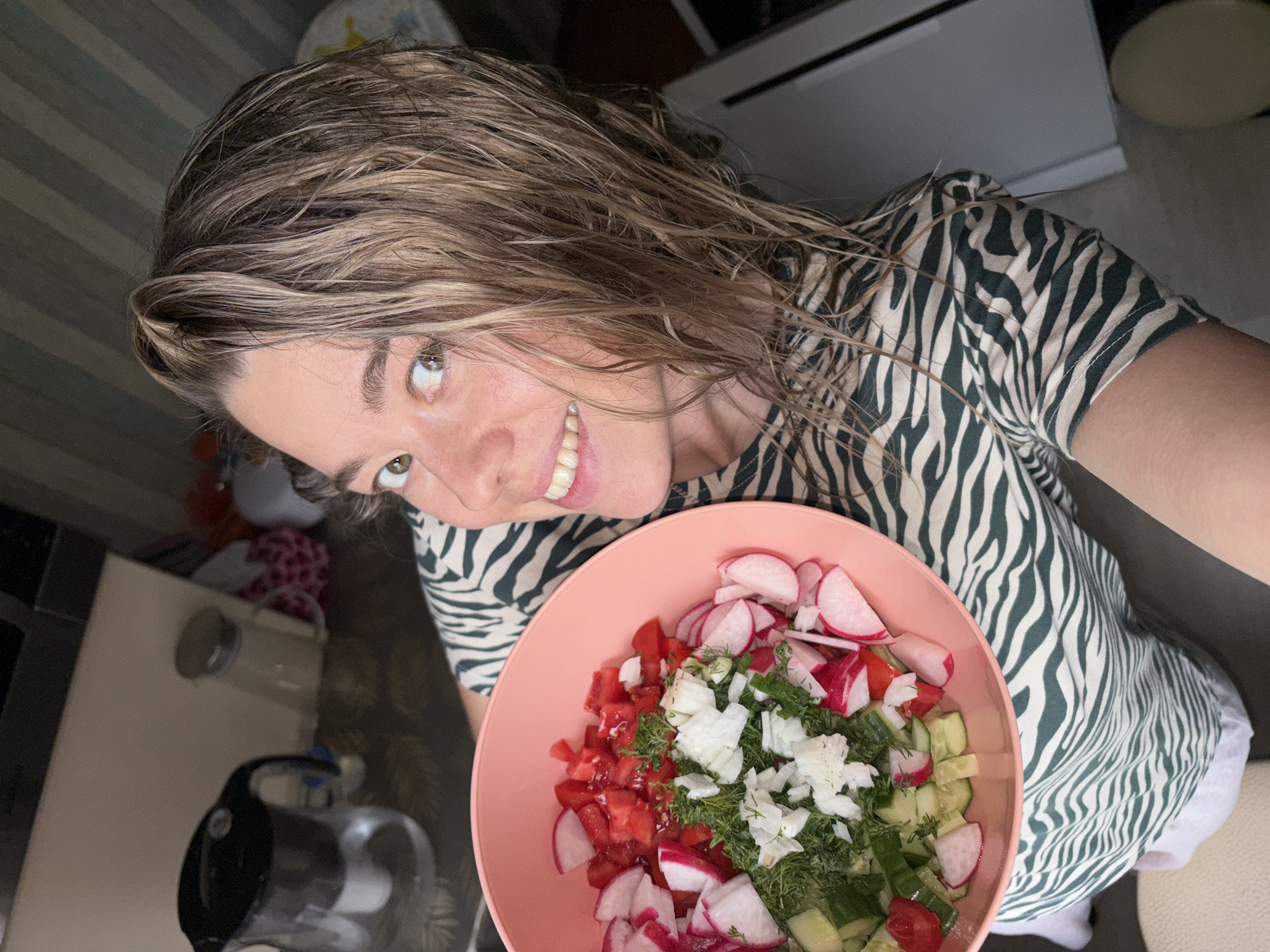 A woman with wet, light-brown hair stands indoors holding a pink bowl of salad. The salad contains diced tomatoes, sliced cucumbers, sliced radishes, chopped onions, and dill. She is wearing a beige and dark green zebra-striped shirt and off-white shorts. In the background are various household appliances. The location is consistent with a domestic kitchen. The city of Chaikovsky, Russia, cannot be confirmed or denied from the provided imagery.