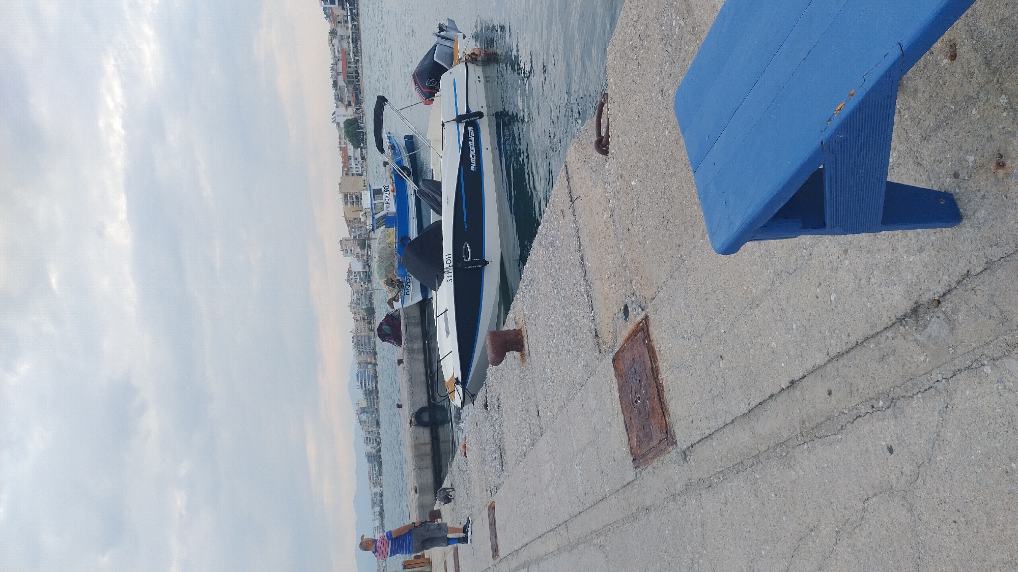 A person stands on a concrete pier next to a small, white and blue motorboat docked in the water. The person, wearing a striped t-shirt and gray shorts, is looking towards the left of the frame and has their left arm raised. A small, gray cat is visible on the pier near the person. The boat, branded "QUICKSILVER," has a black canopy and an outboard motor. In the background, a line of buildings and houses stretches along the coastline under a cloudy sky. Several other boats are moored in the harbor. The scene appears to be in the city of Neoi Epivates, Greece, with the Aegean Sea visible. A bright blue wooden bench is situated in the foreground on the pier.