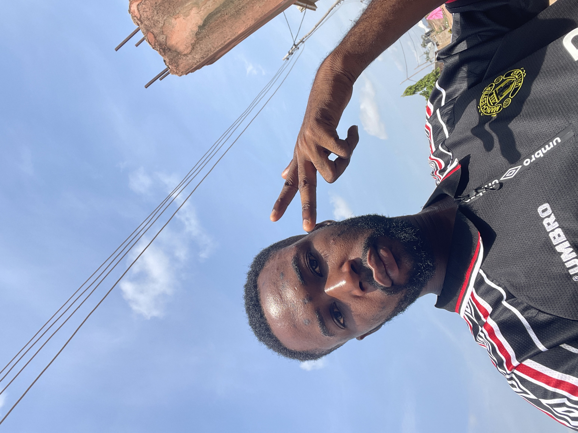 A dark-skinned man with a beard is taking a selfie in Lokoja, Nigeria. He is wearing a black Umbro Manchester United jersey and making a peace sign with his right hand. Overhead power lines are visible in the background against a blue sky with scattered clouds. A partially destroyed concrete structure is in the upper left corner. The location is an urban area in Lokoja, as indicated by the power lines and building structure.