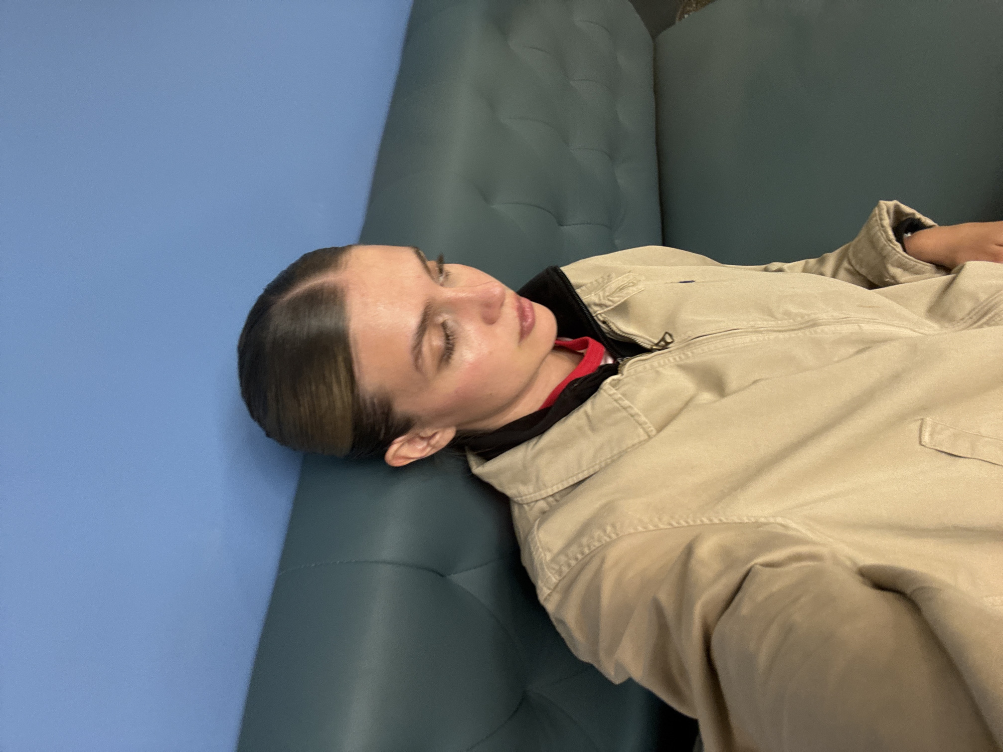 A person is lying on a light blue-grey upholstered couch or seating area against a light blue wall. The person appears to be resting with eyes closed. They are wearing a light tan jacket with a dark neckline revealing a red garment beneath. Their hair is dark and pulled back, with some lighter highlights visible. The location is presumed to be in Tbilisi, Georgia.